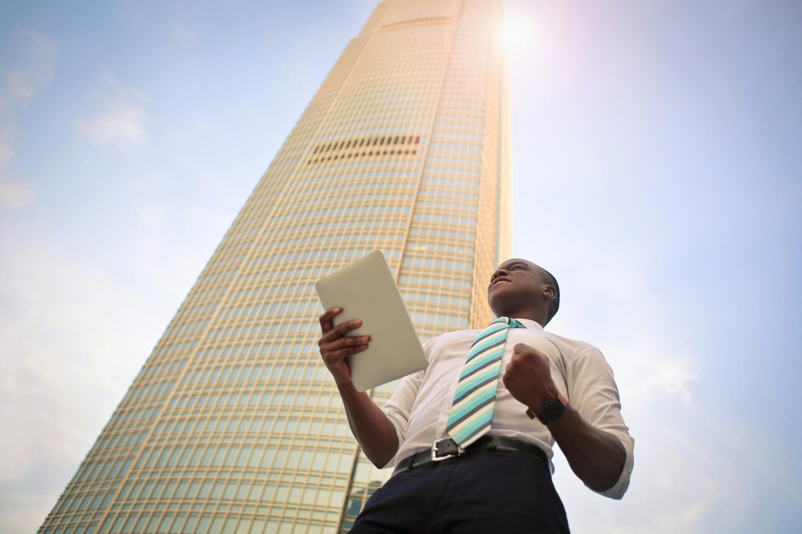 man standing outside skyscraper building wearing smart trousers, shirt and tie and holding a tablet