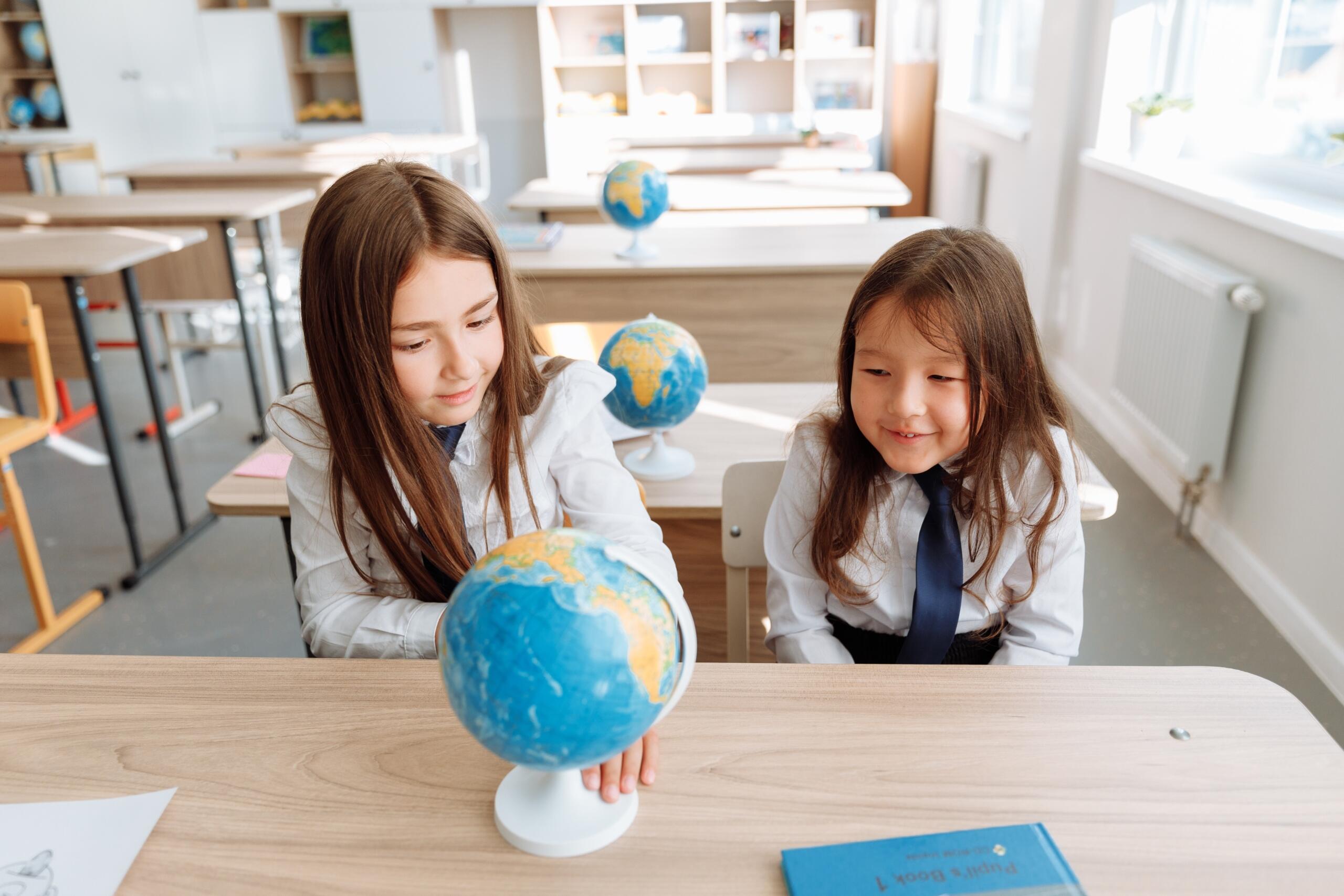 two young girls wearing school uniforms sitting in a classroom looking at a map of the world