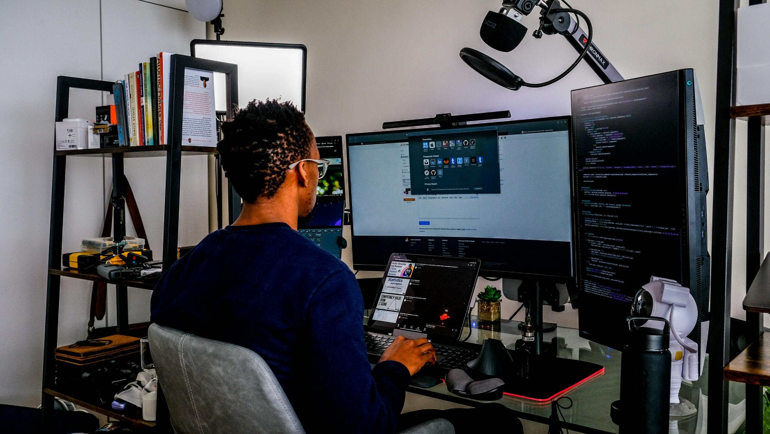 man sitting at desk in front of laptop and multiple computer monitors coding