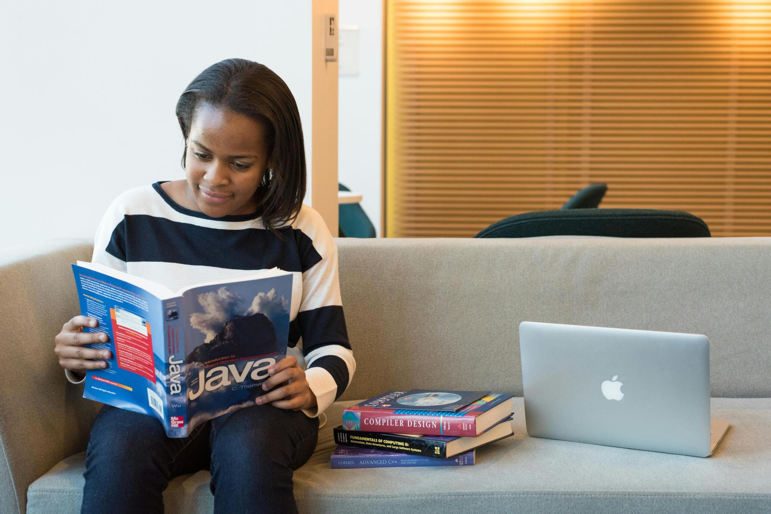 person sitting on sofa next to laptop and a pile of books reading book about Java coding