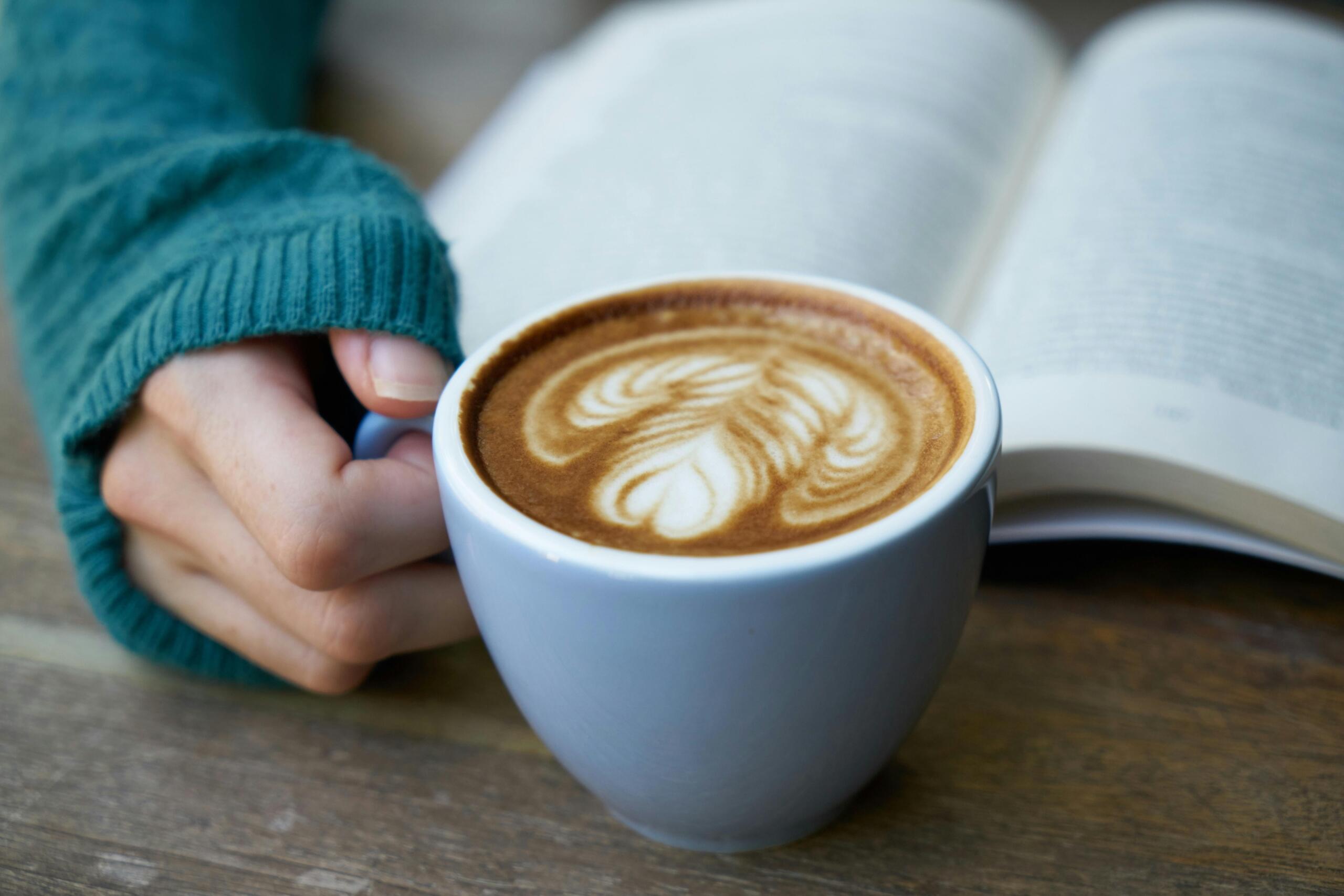 woman holding coffee cup while reading a book placed on a wooden table