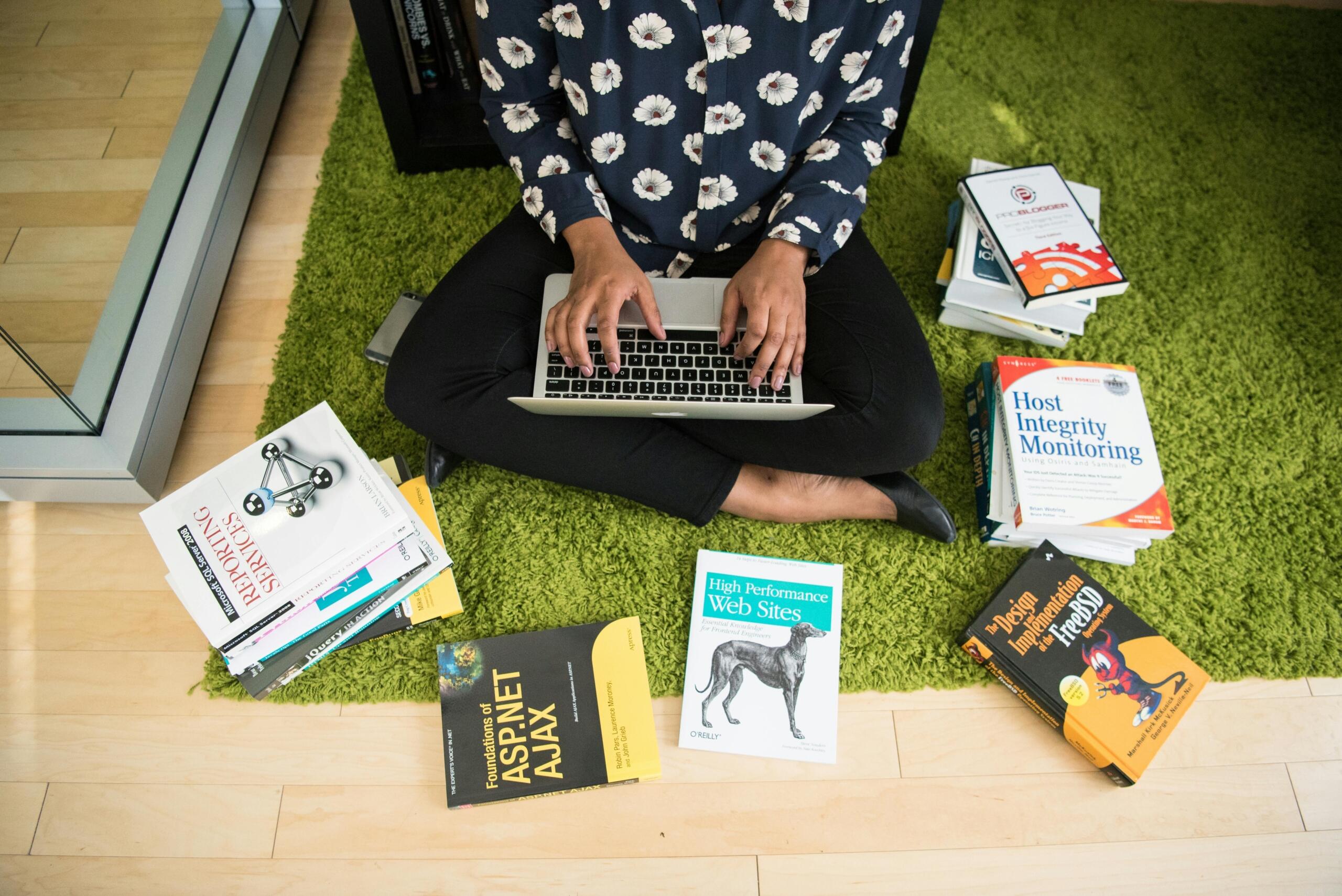 woman sitting on floor surrounded by coding books and using laptop