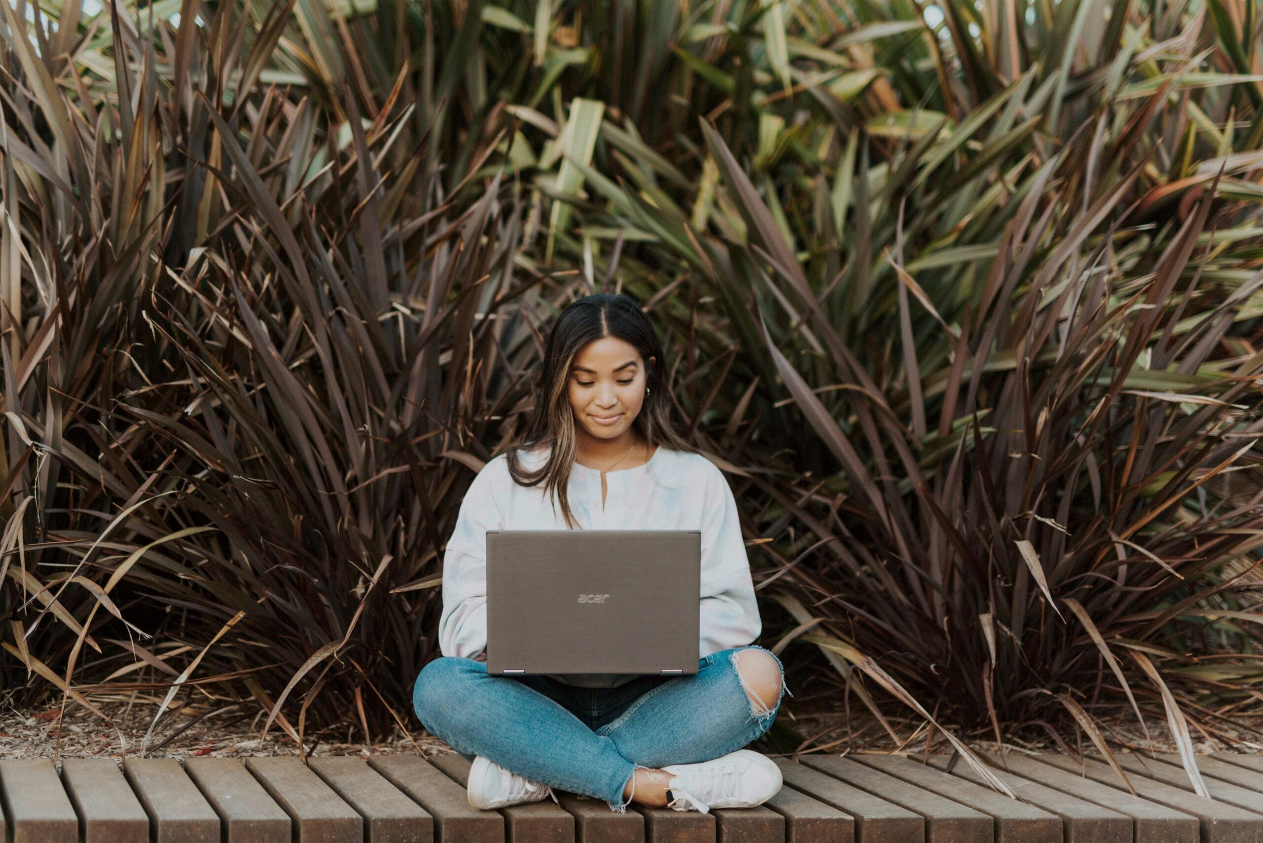 young woman sitting cross-legged outside on wooden walkway working on a laptop
