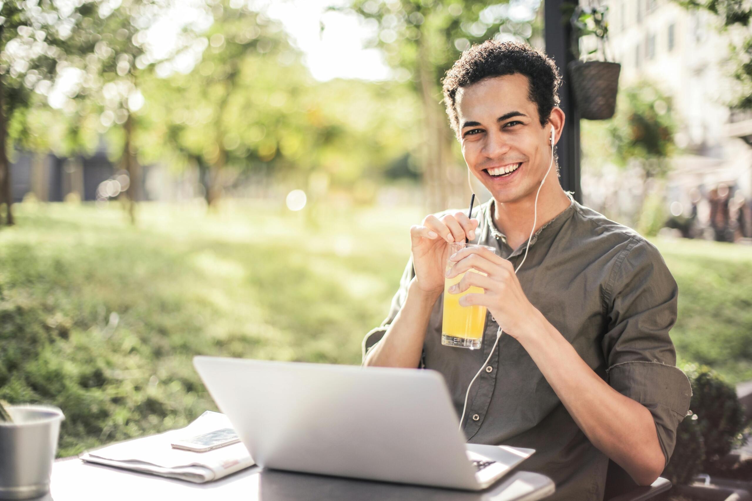 happy man wearing earphones sitting with laptop and juice in park