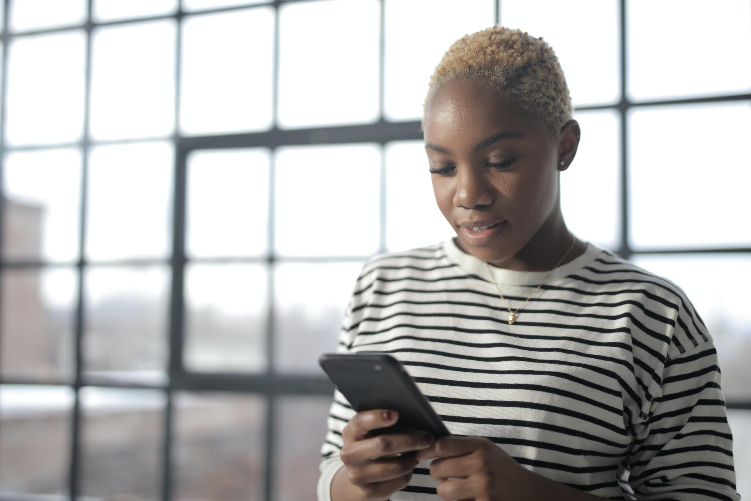 woman standing in office with large black-framed windows looking at mobile phone