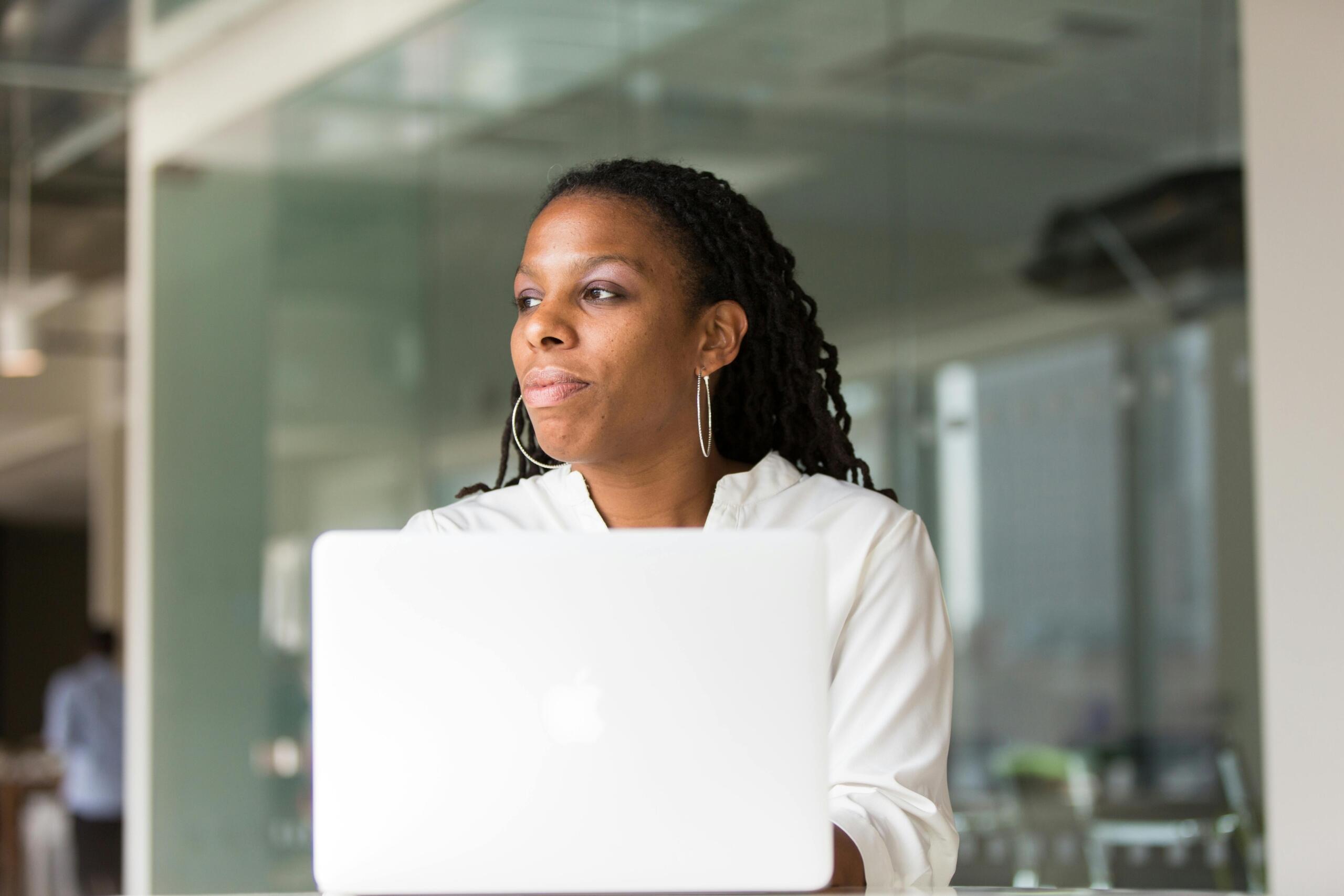 woman staring into the distance seated in office using laptop
