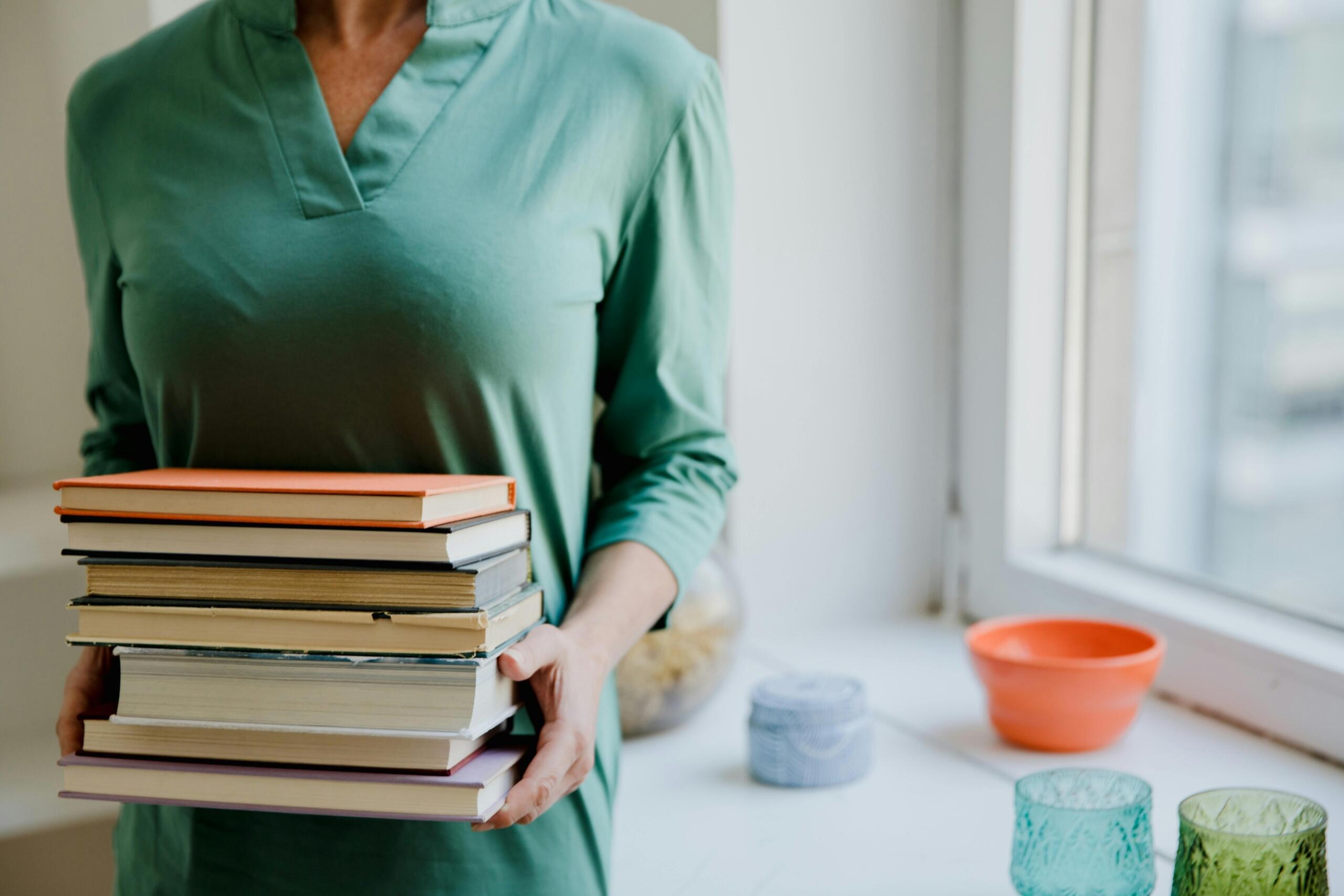 woman standing next to a window holding a large pile of books in her hands