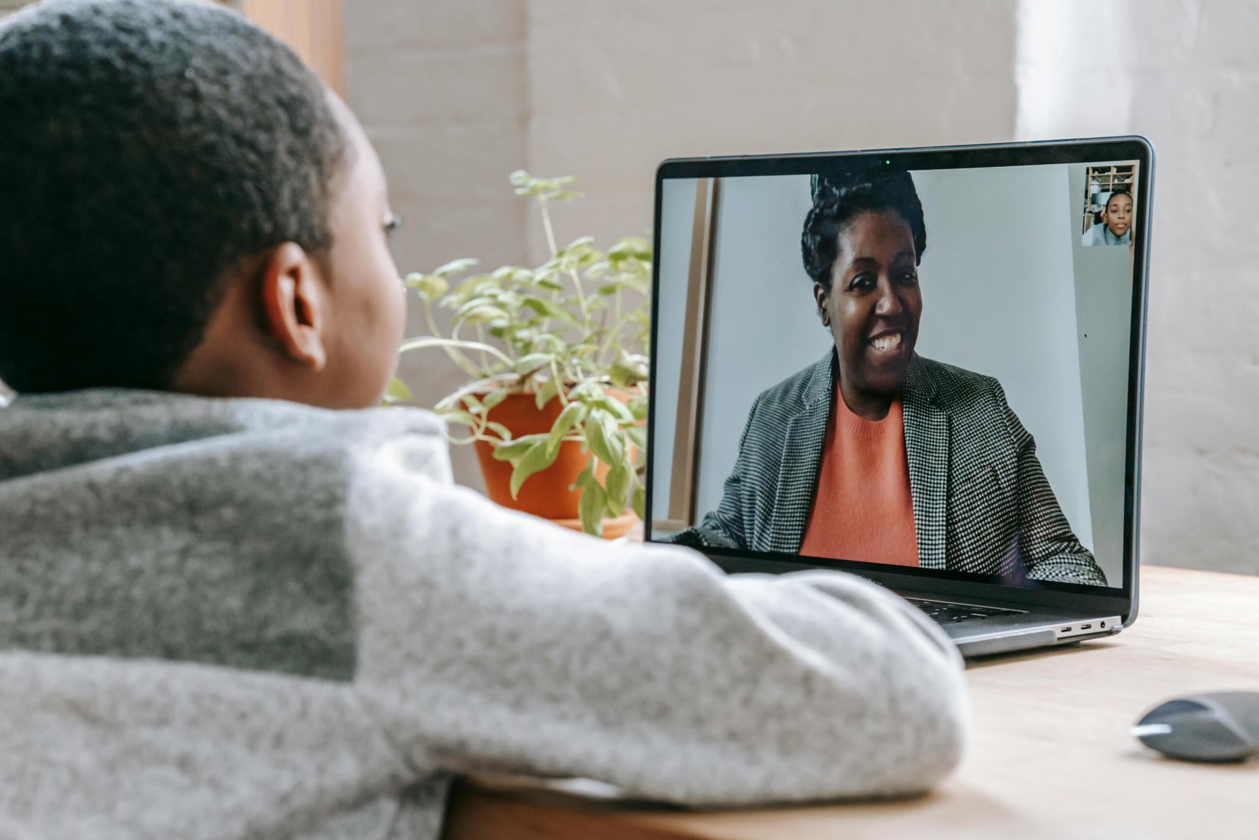 boy sitting at desk connected to online class with teacher on laptop