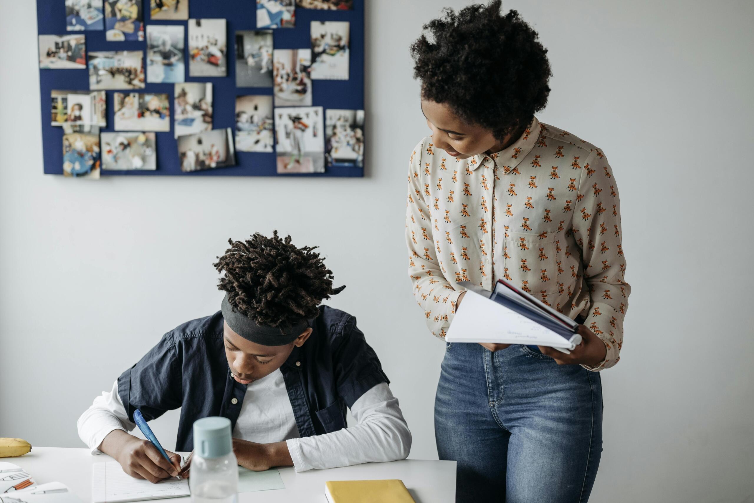 teacher standing next to student in class watching as he completes a task in his notebook