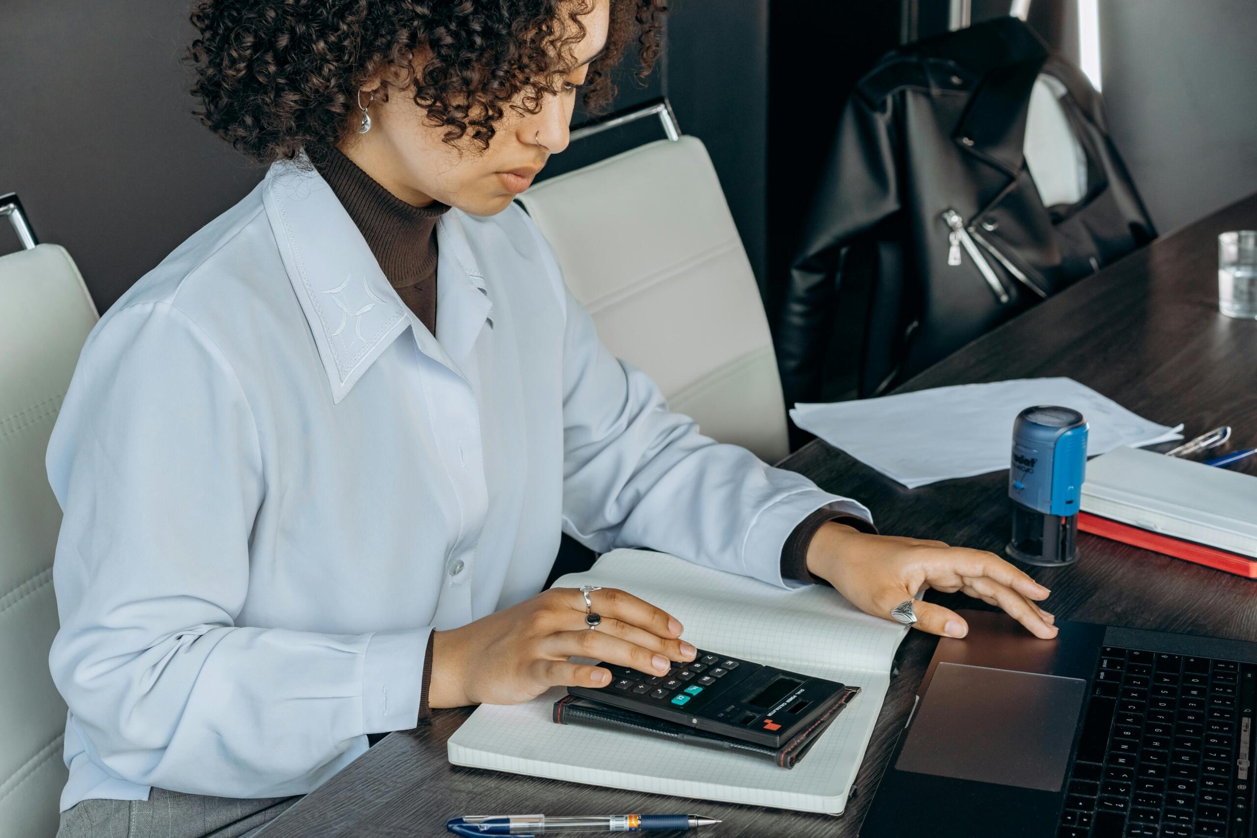 woman wearing professional clothing sitting at a desk typing on a laptop and using a calculator