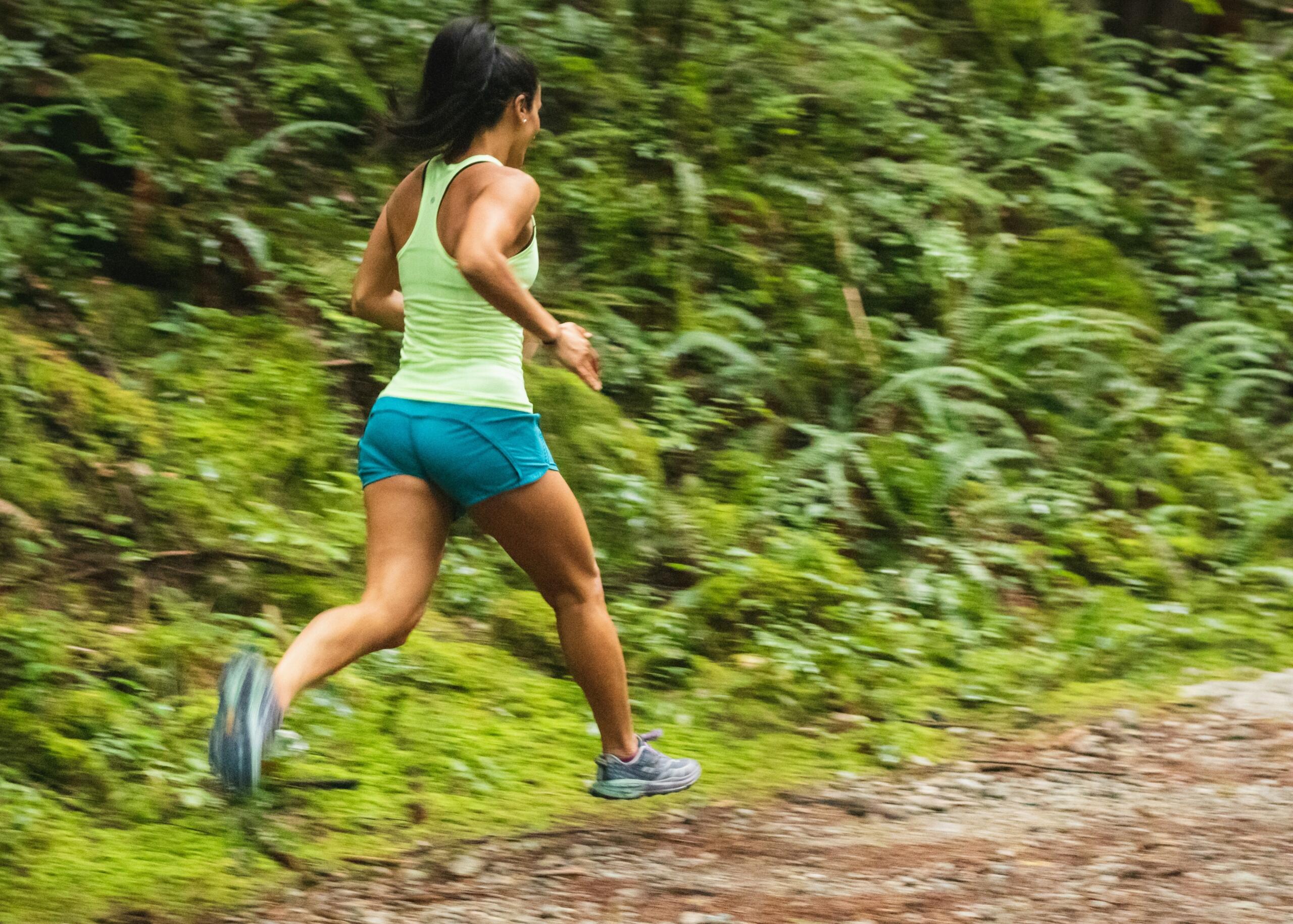 woman wearing neon green running vest and blue shorts running on trail in the forest