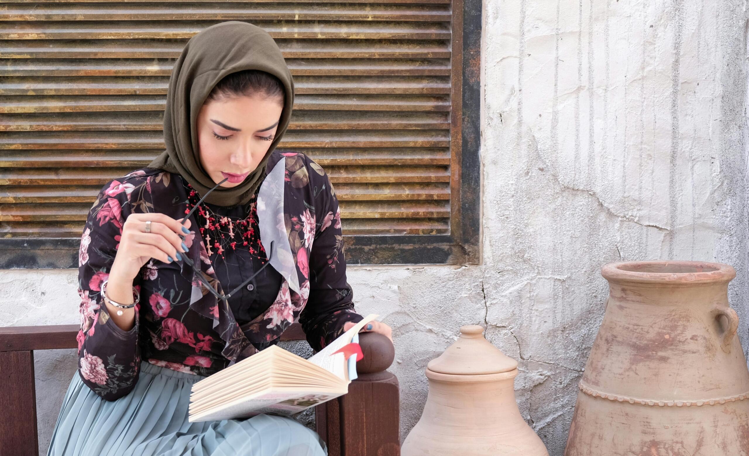 woman wearing headscarf and holding spectacles in her hand while reading a book