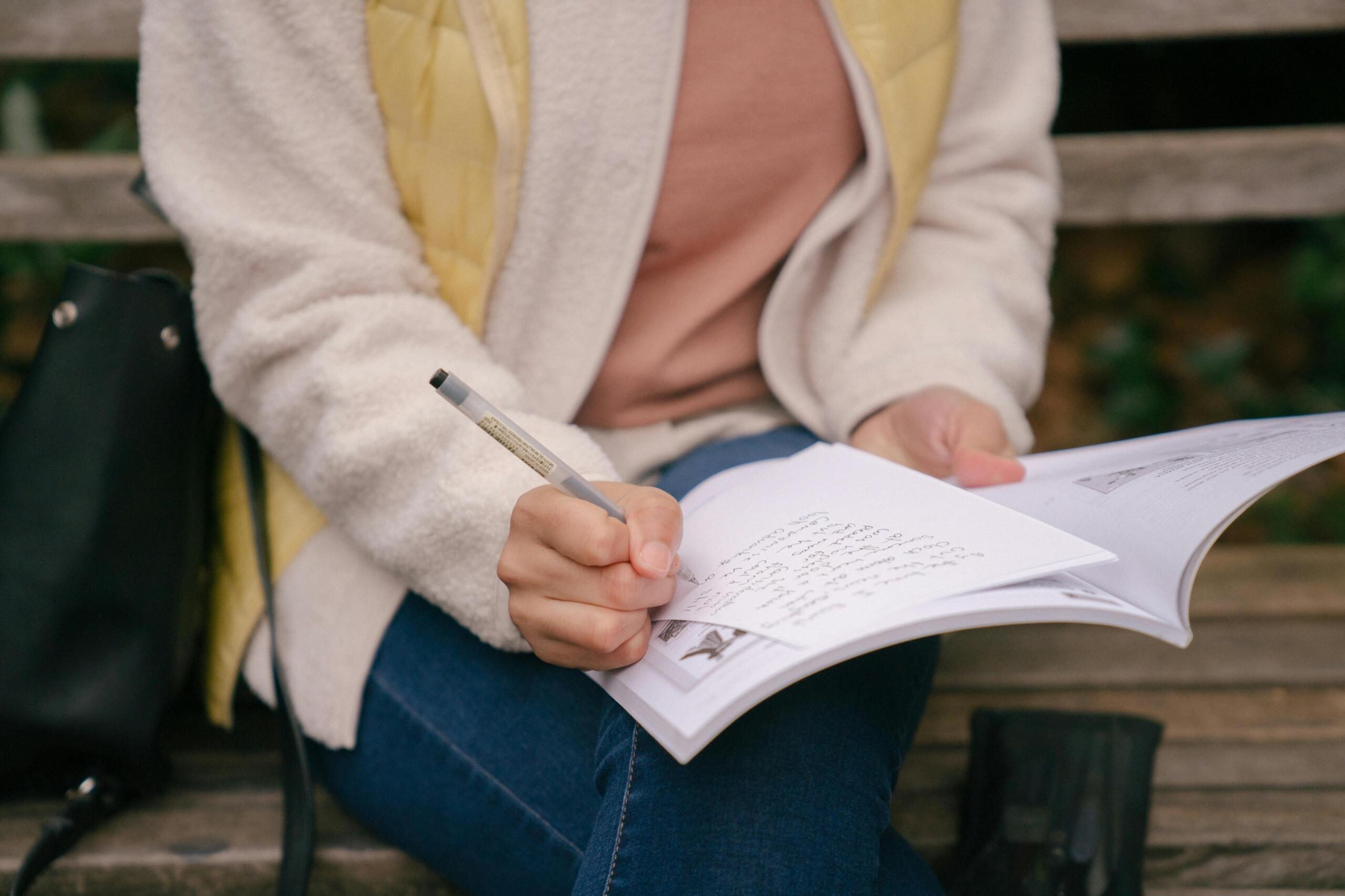 person writing in a notebook sitting on a bench with a black sling bag alongside them