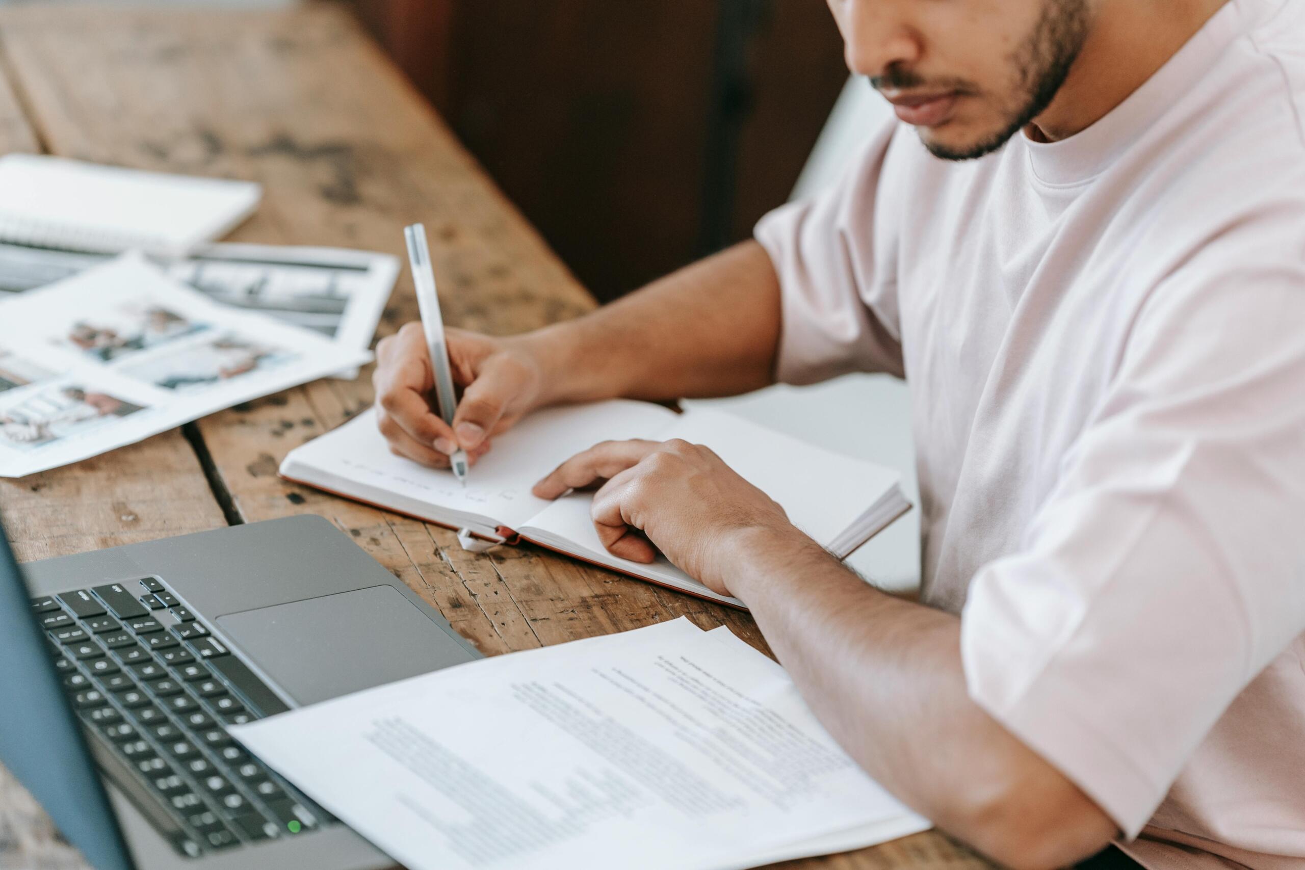 young man making notes and reading from printouts, while using a laptop