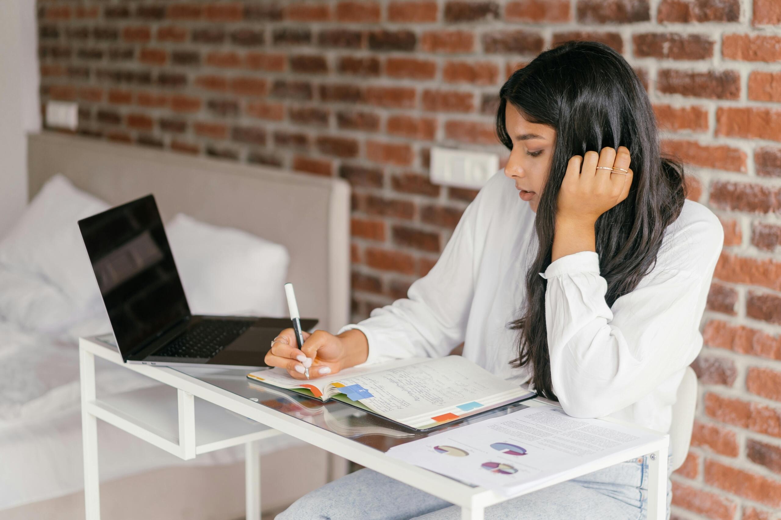 girl sitting at a small desk writing in a notebook and using a laptop