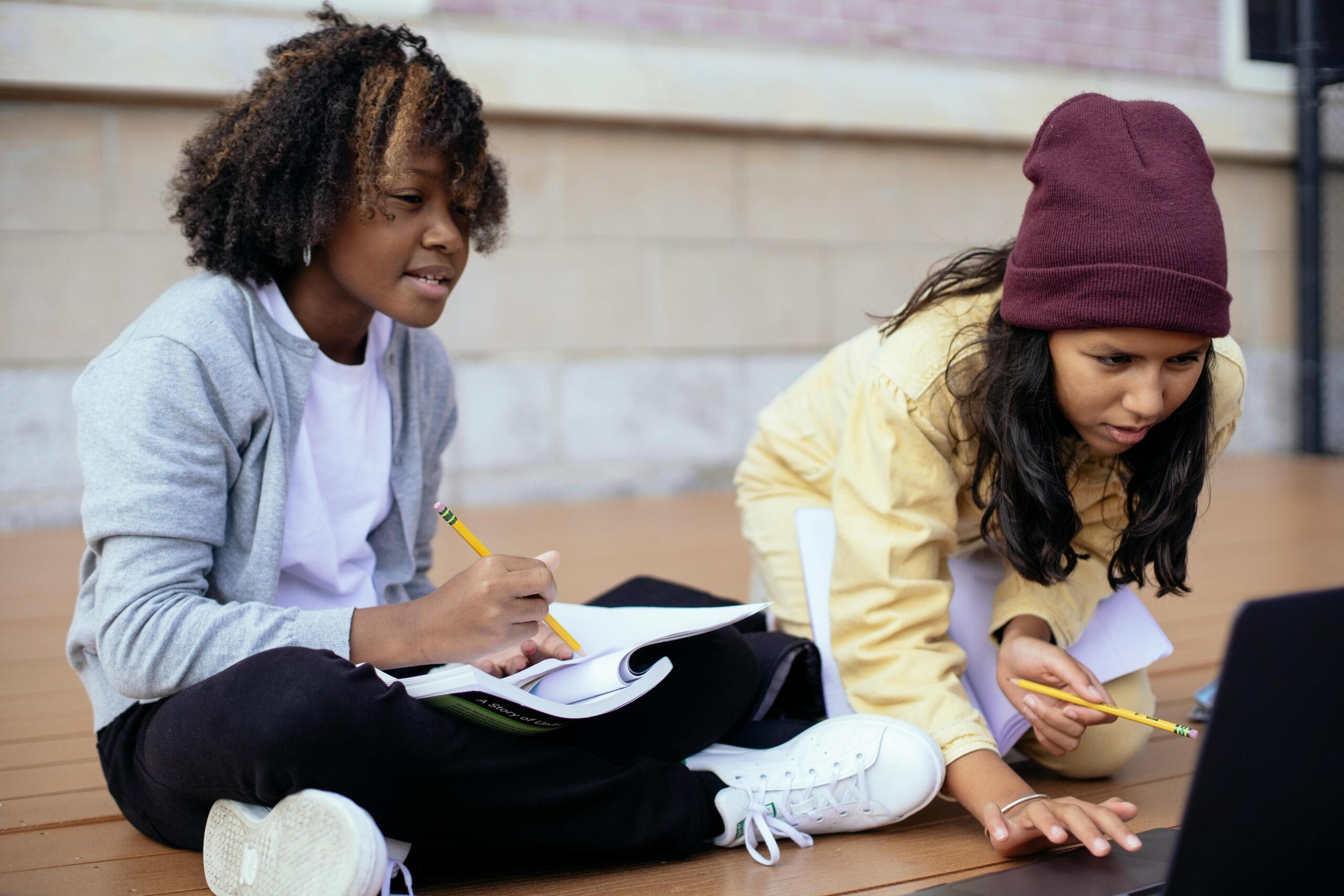 two schoolgirls outside using laptop and writing in notebooks