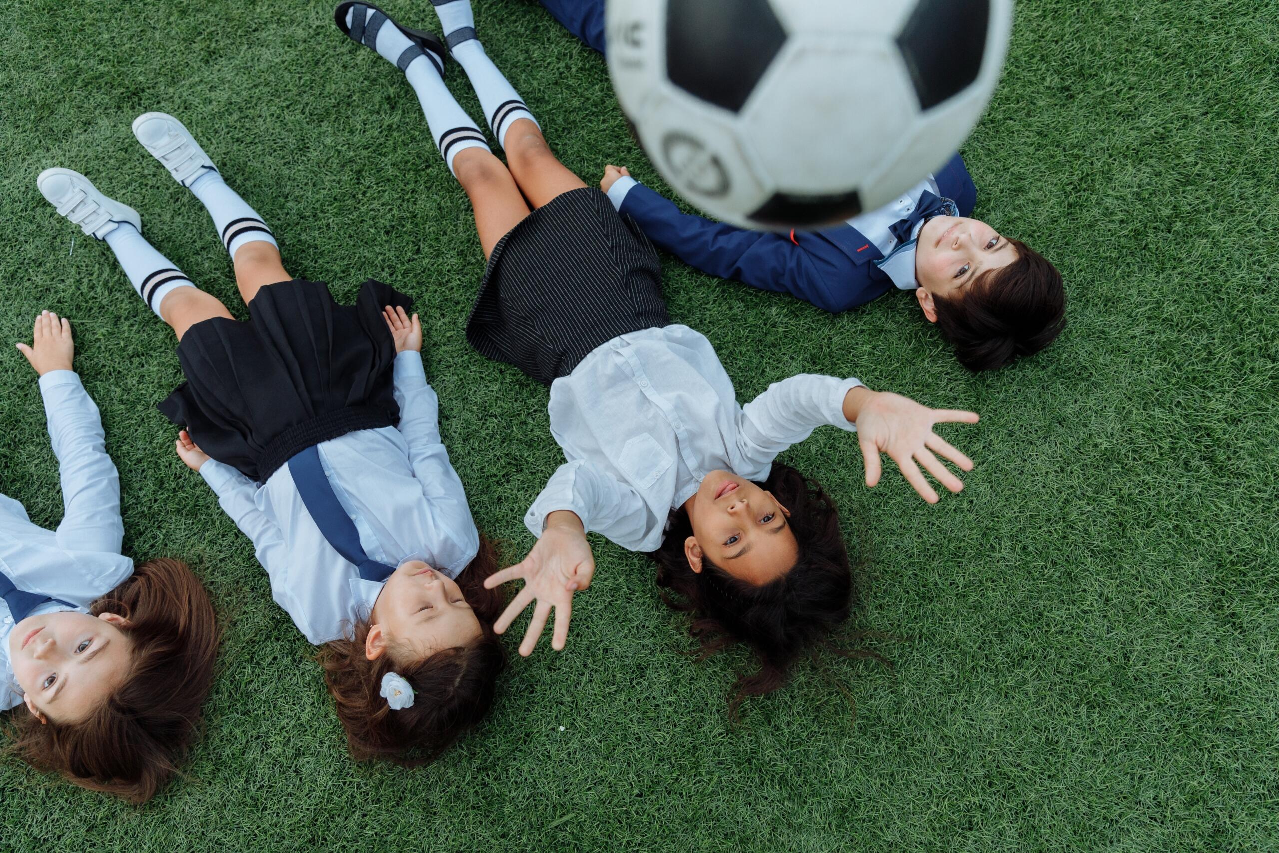four school children lying on astroturf throwing a soccer ball in the air