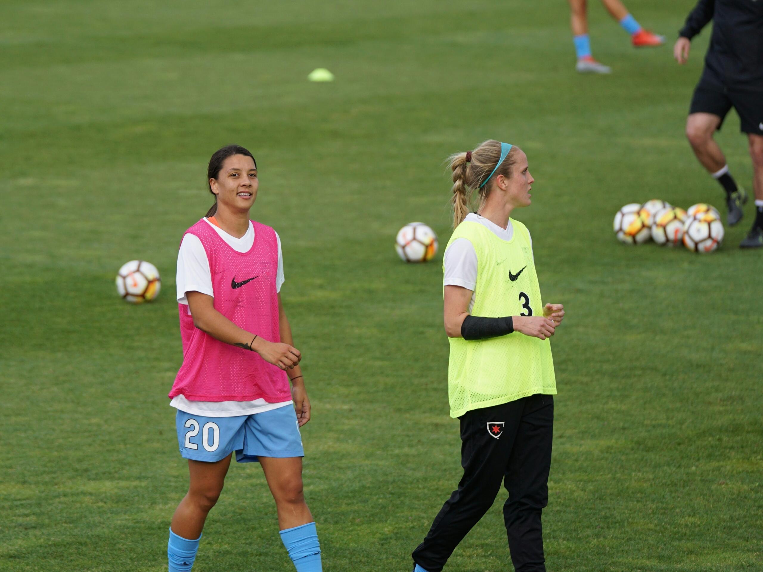 two young woman in soccer training kits on a soccer field with soccer balls in the background