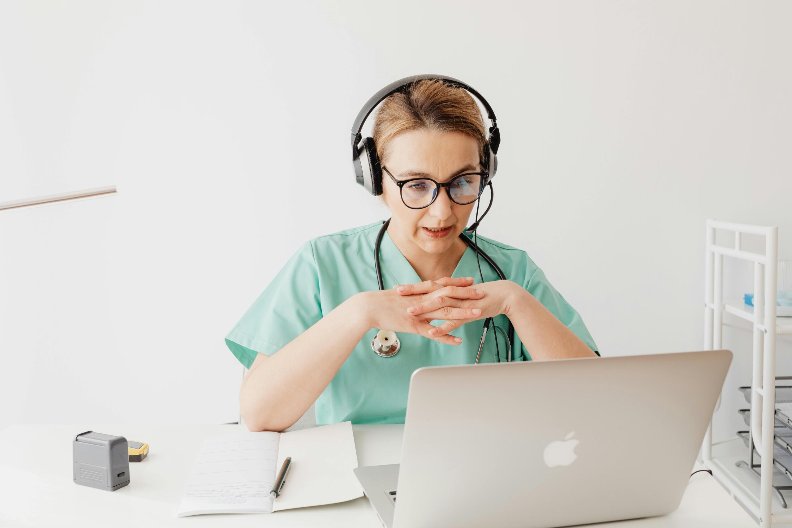 doctor in green scrubs with stethoscope draped over her shoulders, wearing headphones, sitting in front of a laptop