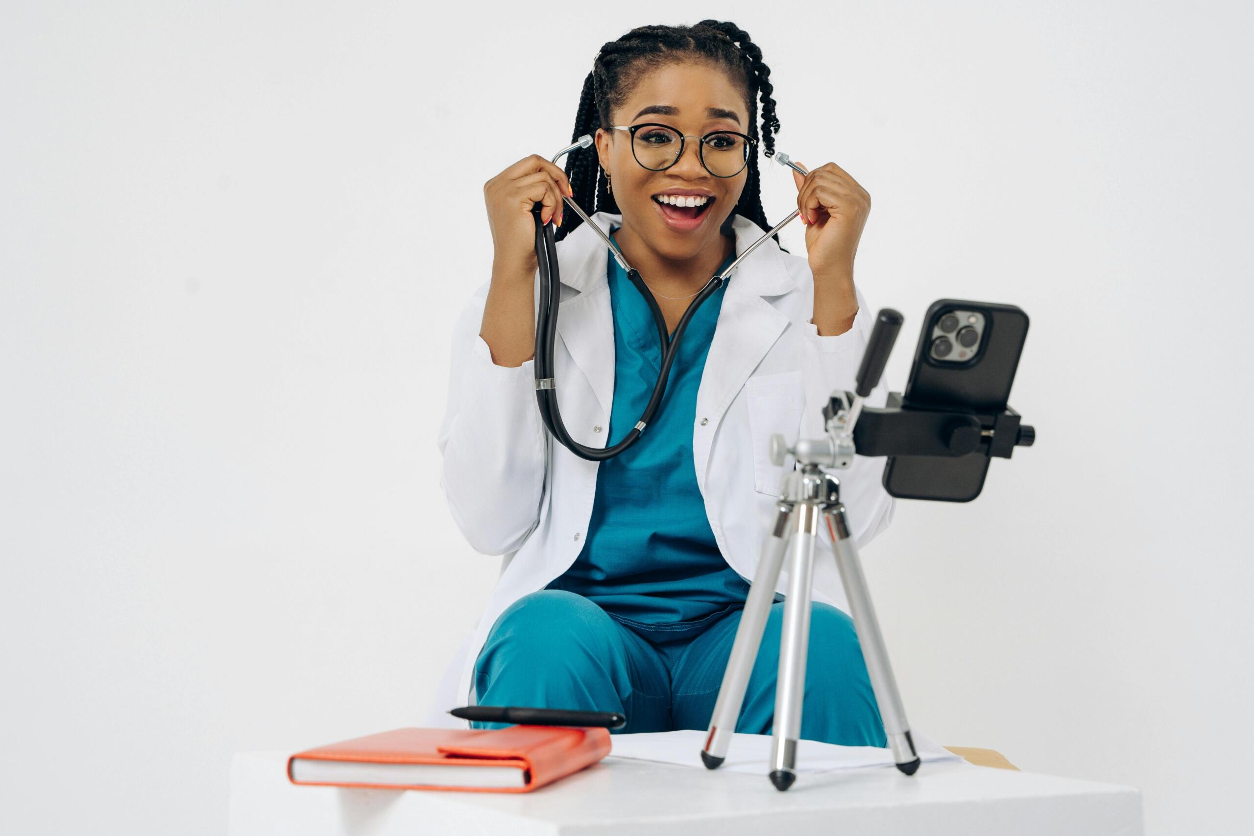 doctor in blue scrubs holding stethoscope up to her ears sitting in front of a phone balanced on a tripod on a table next to a notebook
