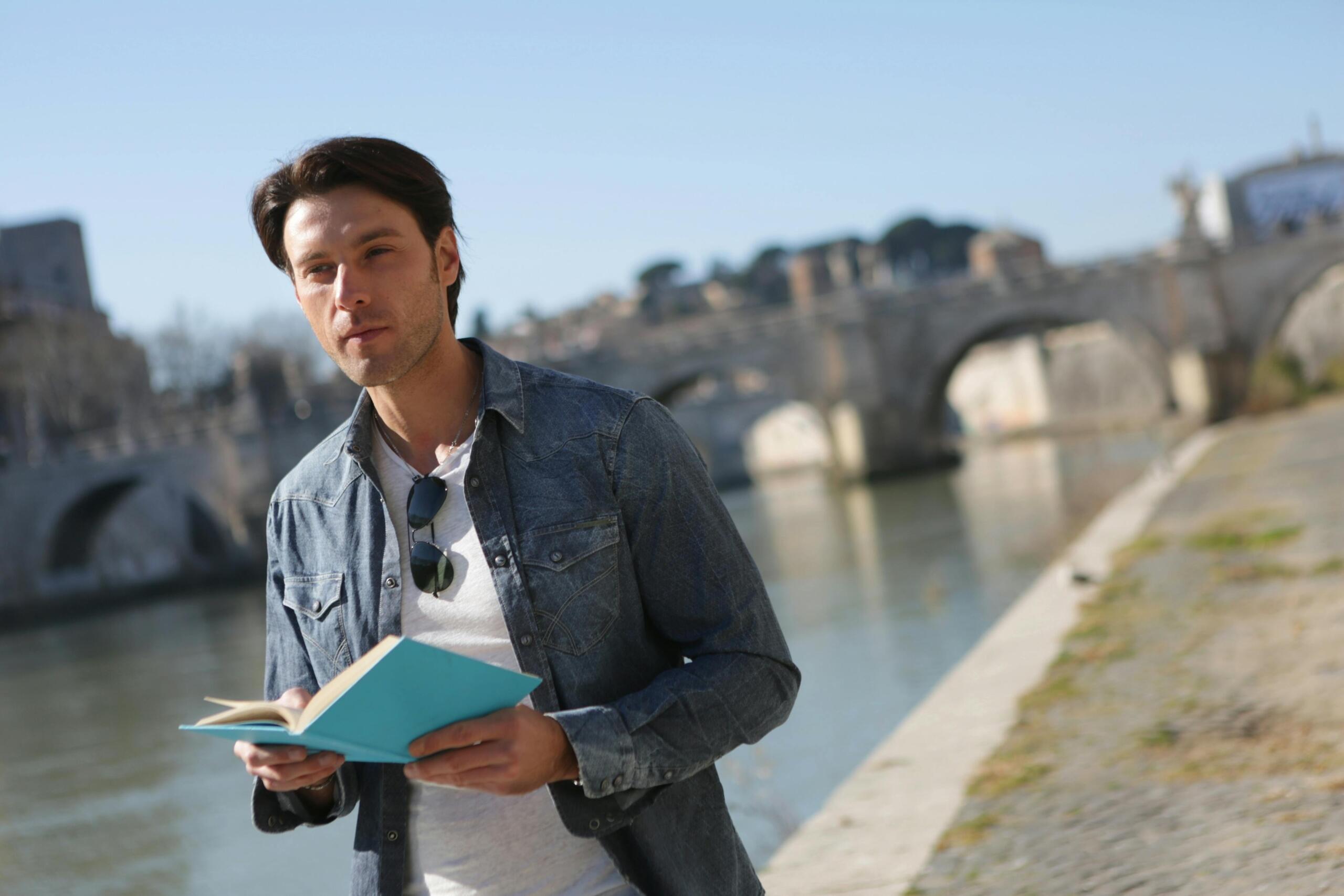 man walking on the banks of a river near a stone bridge holding a book