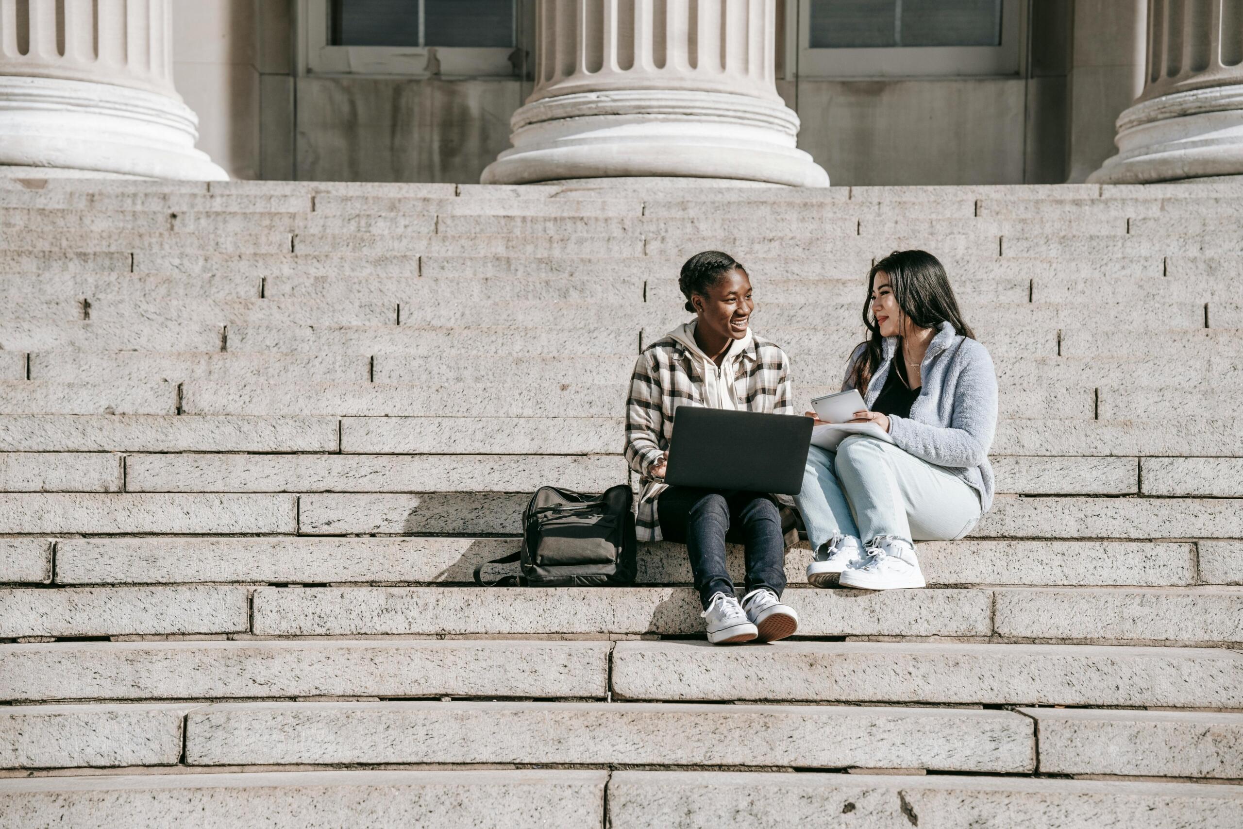 two young women sitting on stone steps outside a building surrounded by pillars