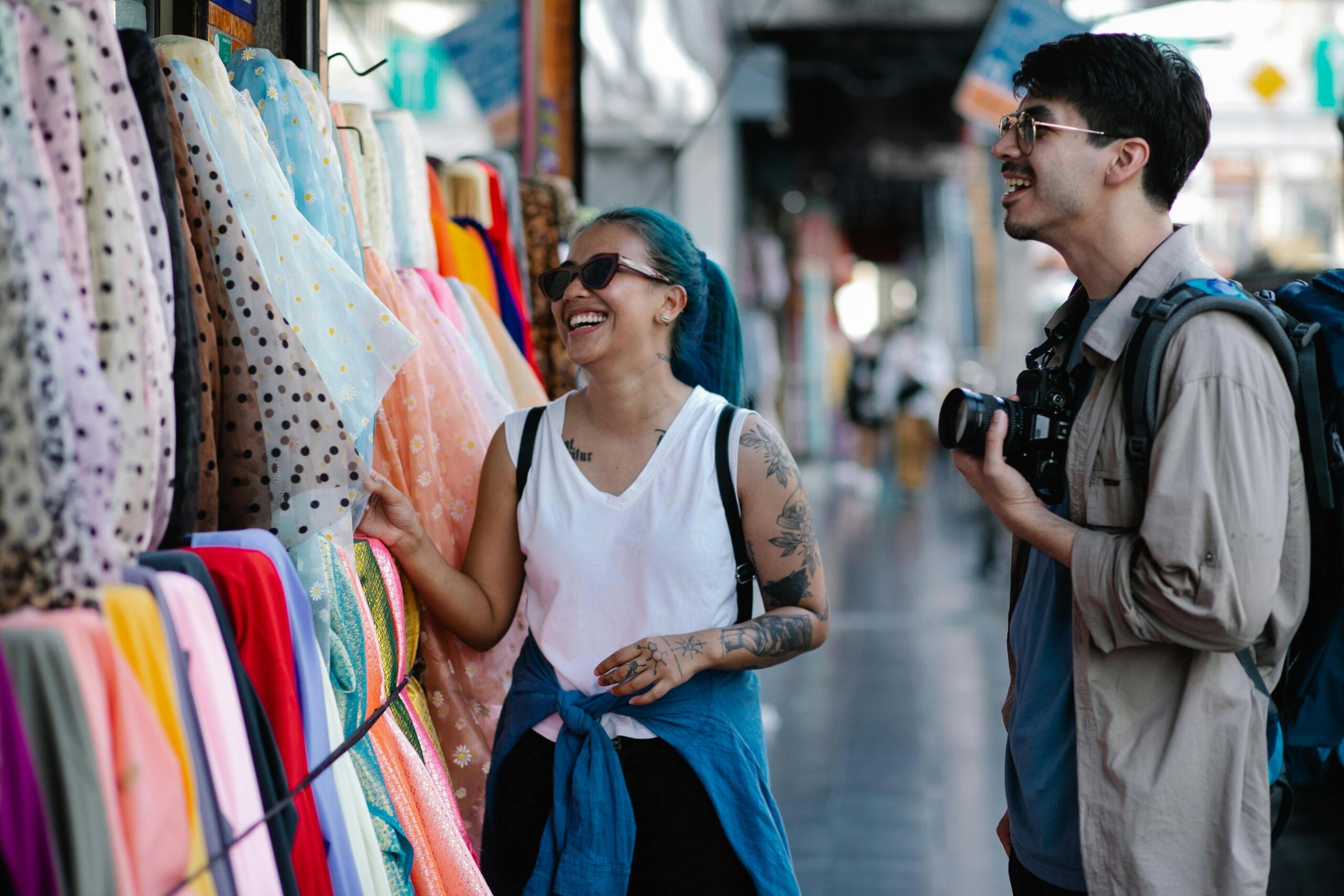 a man holding a camera while a woman touches fabric displayed on the sidewalk in front of a shop