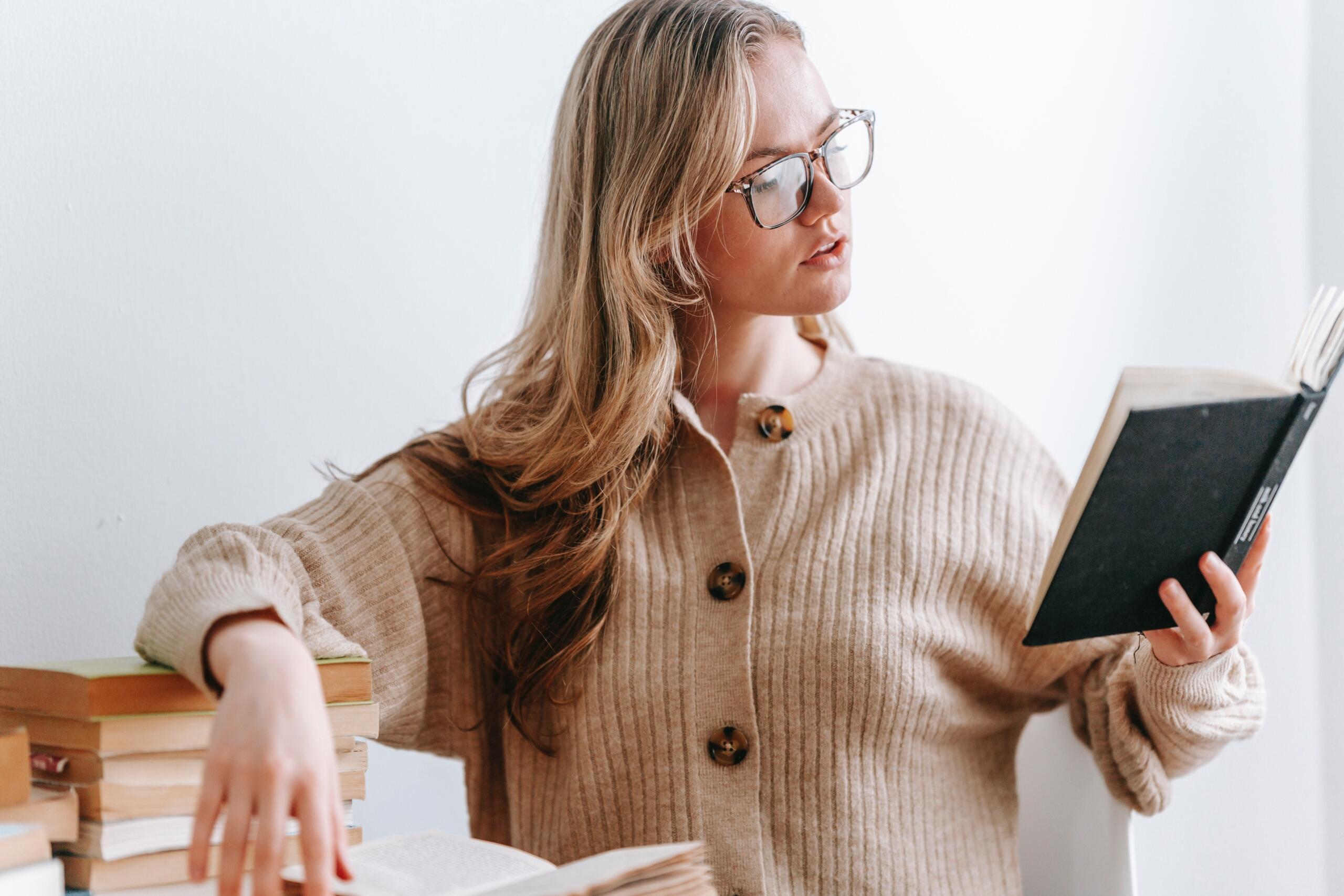 woman standing up leaning on a pile of books, and holding a book in her hand