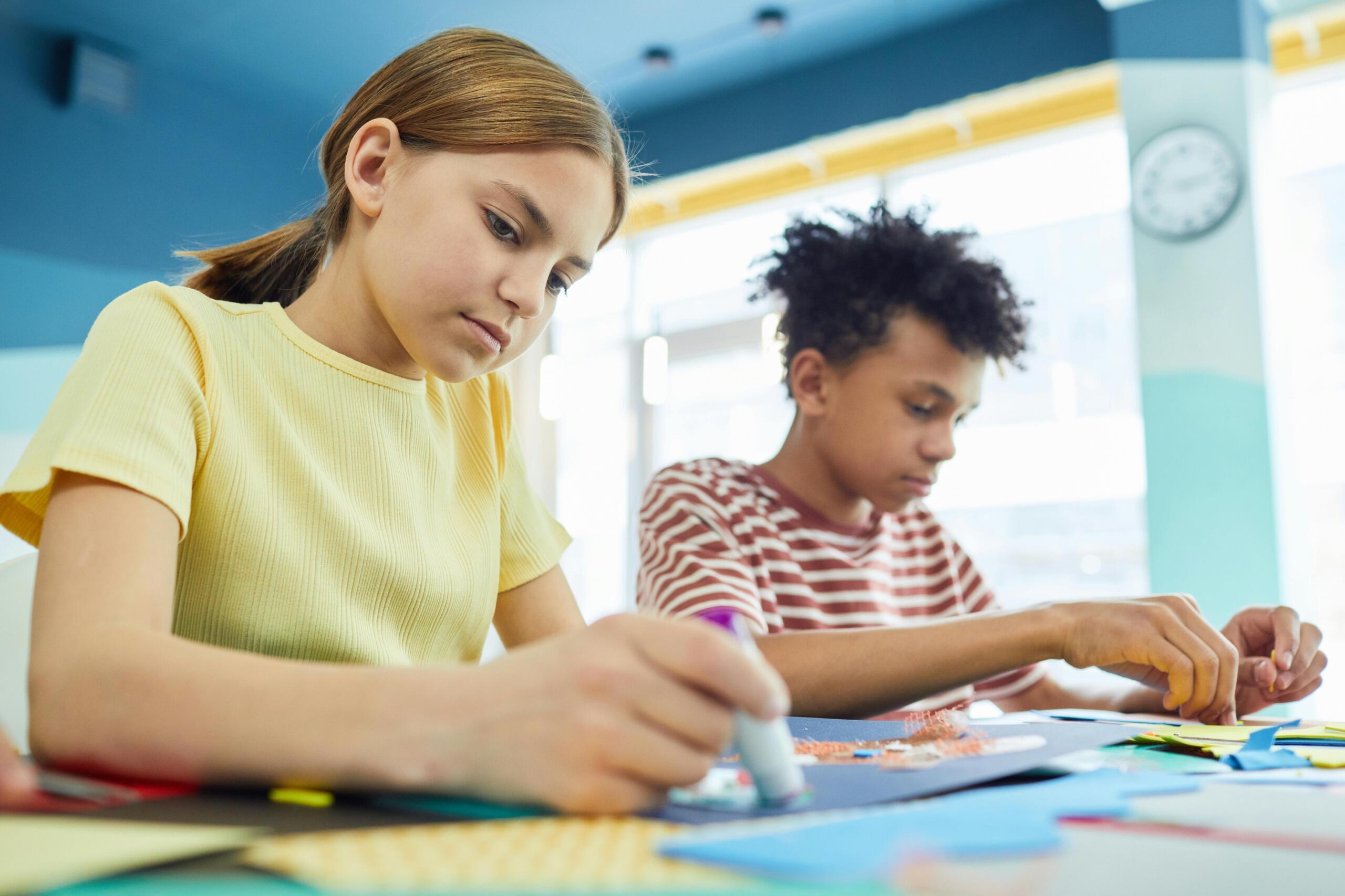 a boy and a girl sitting at a desk glueing items to coloured sheets of paper