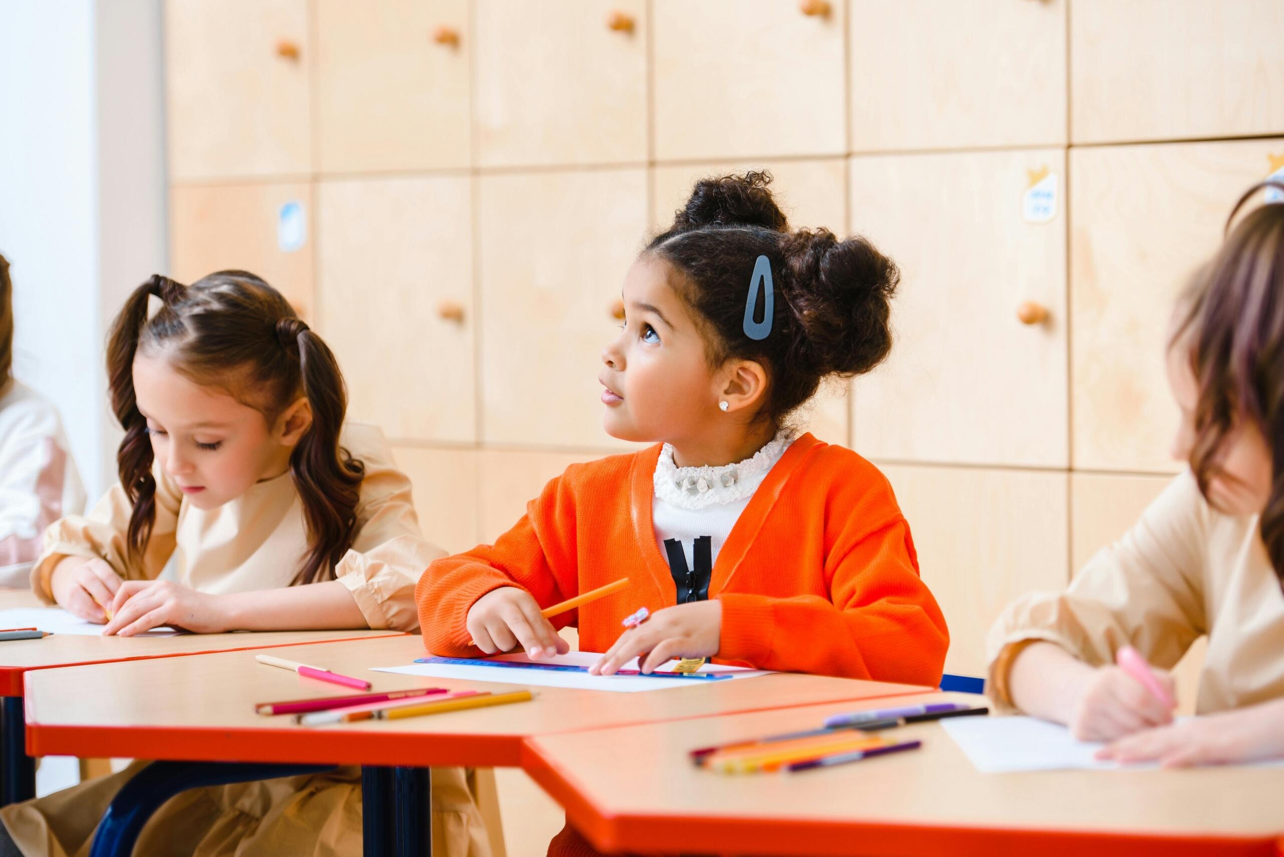 young girl sitting at a desk using a ruler and coloured pencil looking up at teacher