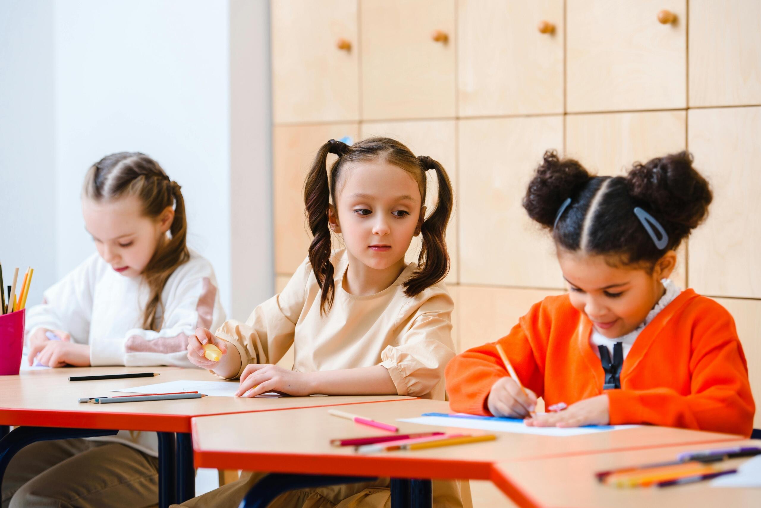 three young girls sitting at desks using coloured pencils