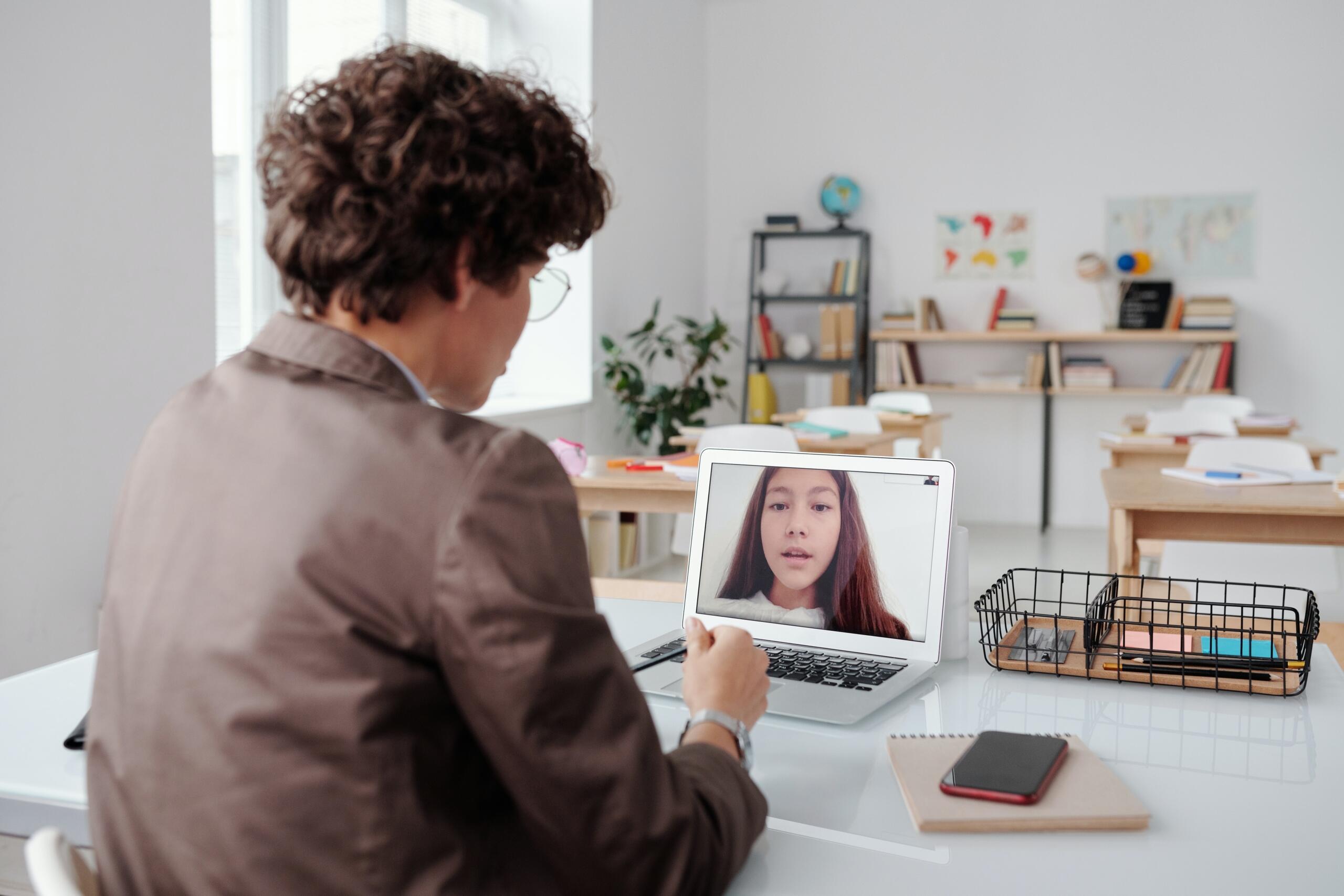 teacher sitting behind desk in classroom on a laptop video call with a student