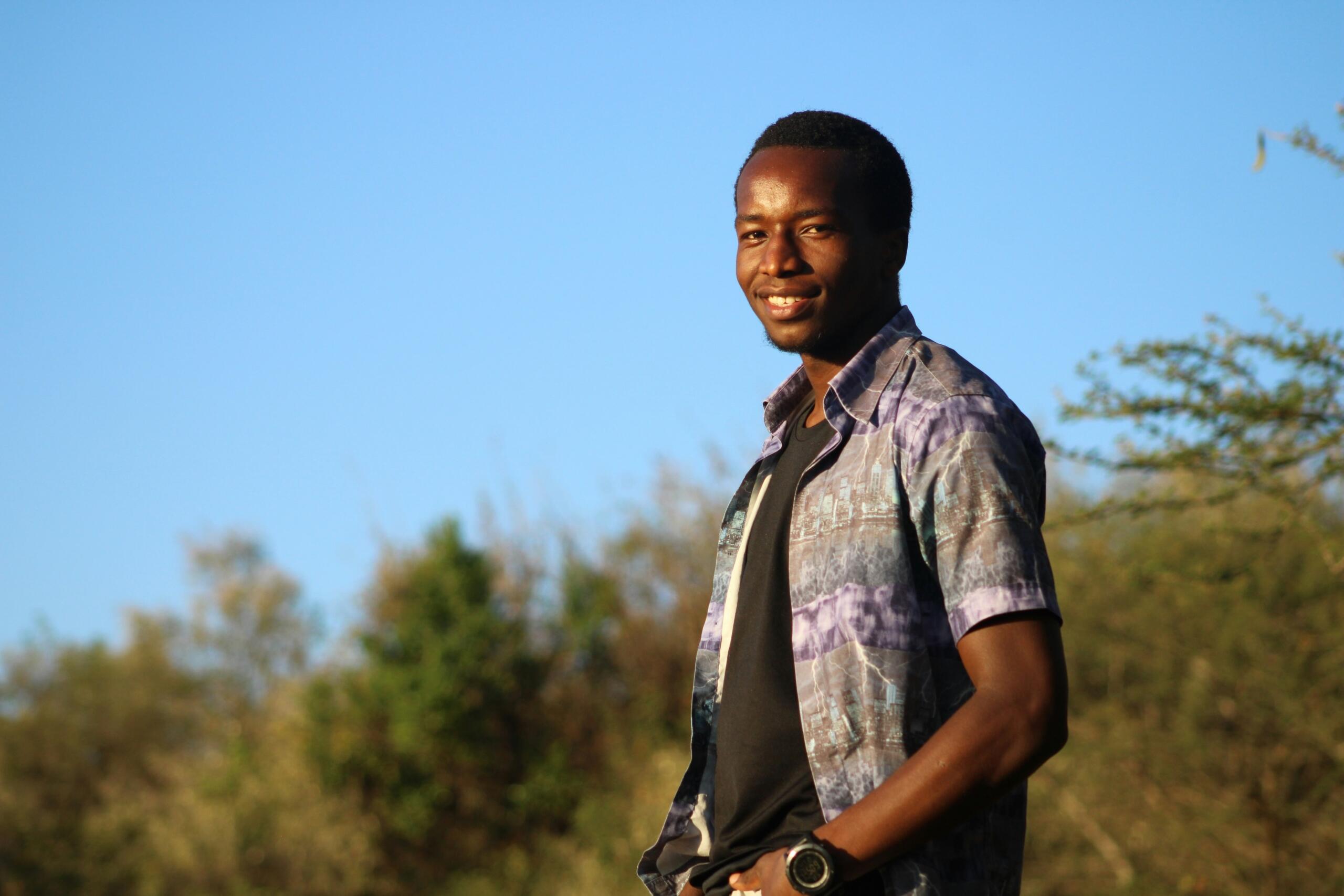 A young man stands against a natural landscape.