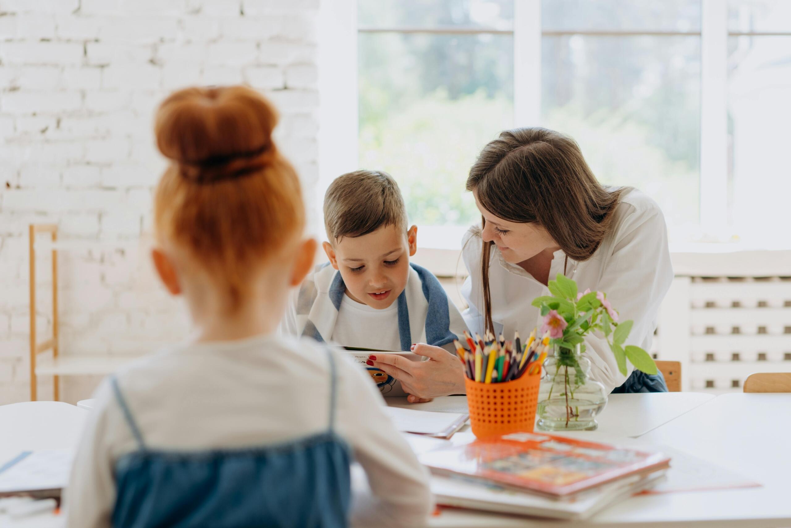 mom sitting at a table helping her son read while daughter reads on her own