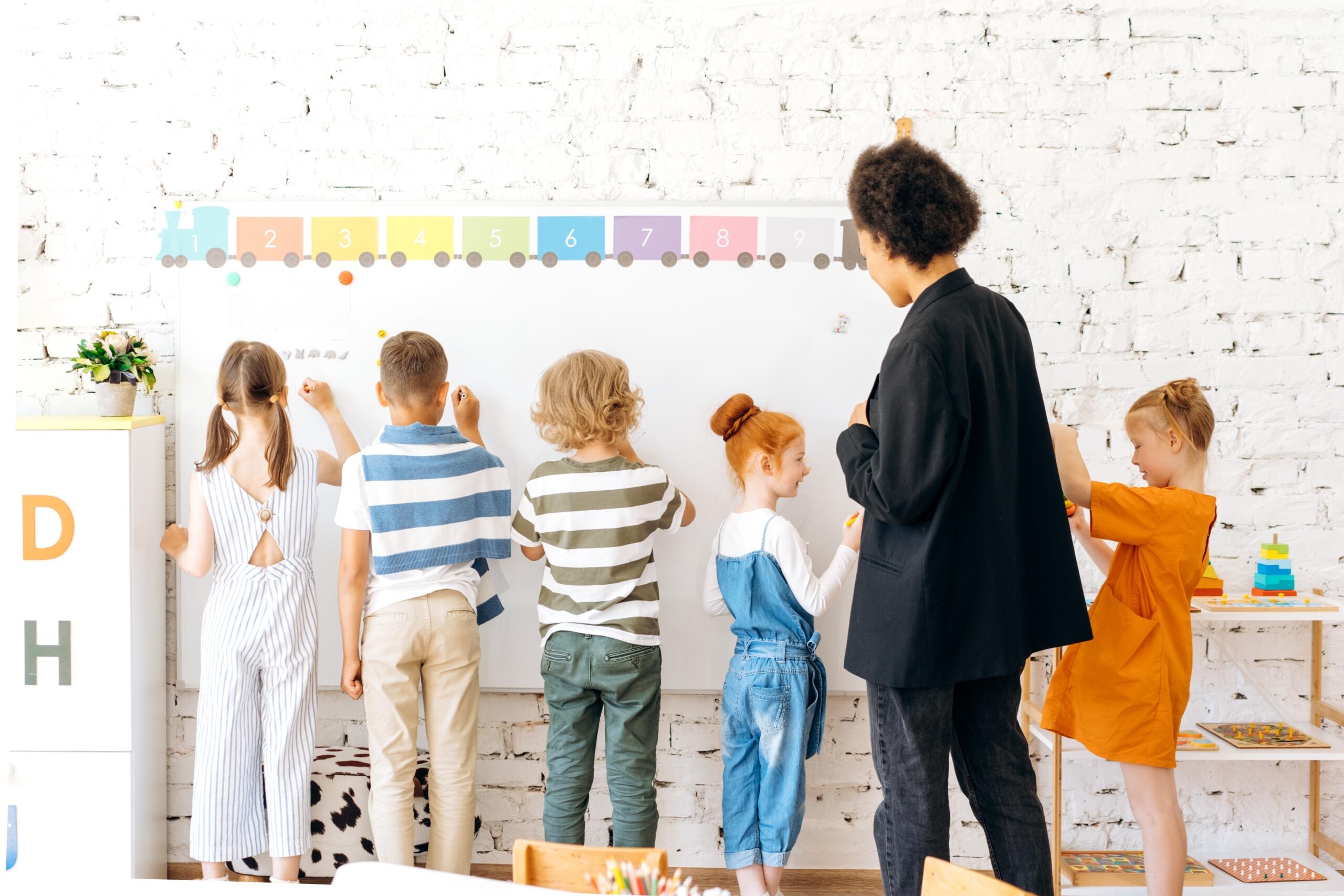 a group of school children standing at the whiteboard in class writing as teacher looks on