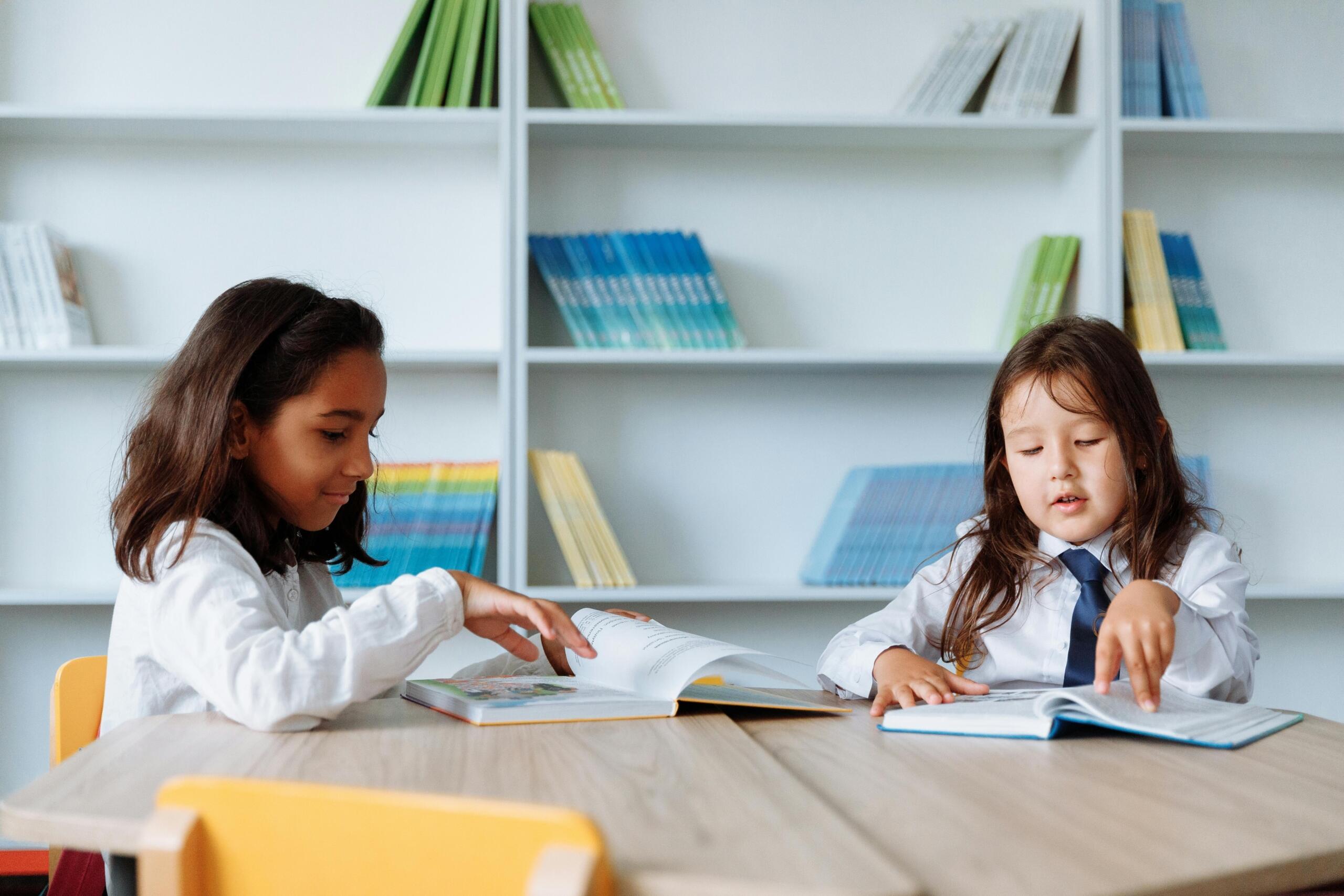 two girls wearing school uniforms sitting at a table in a library reading books