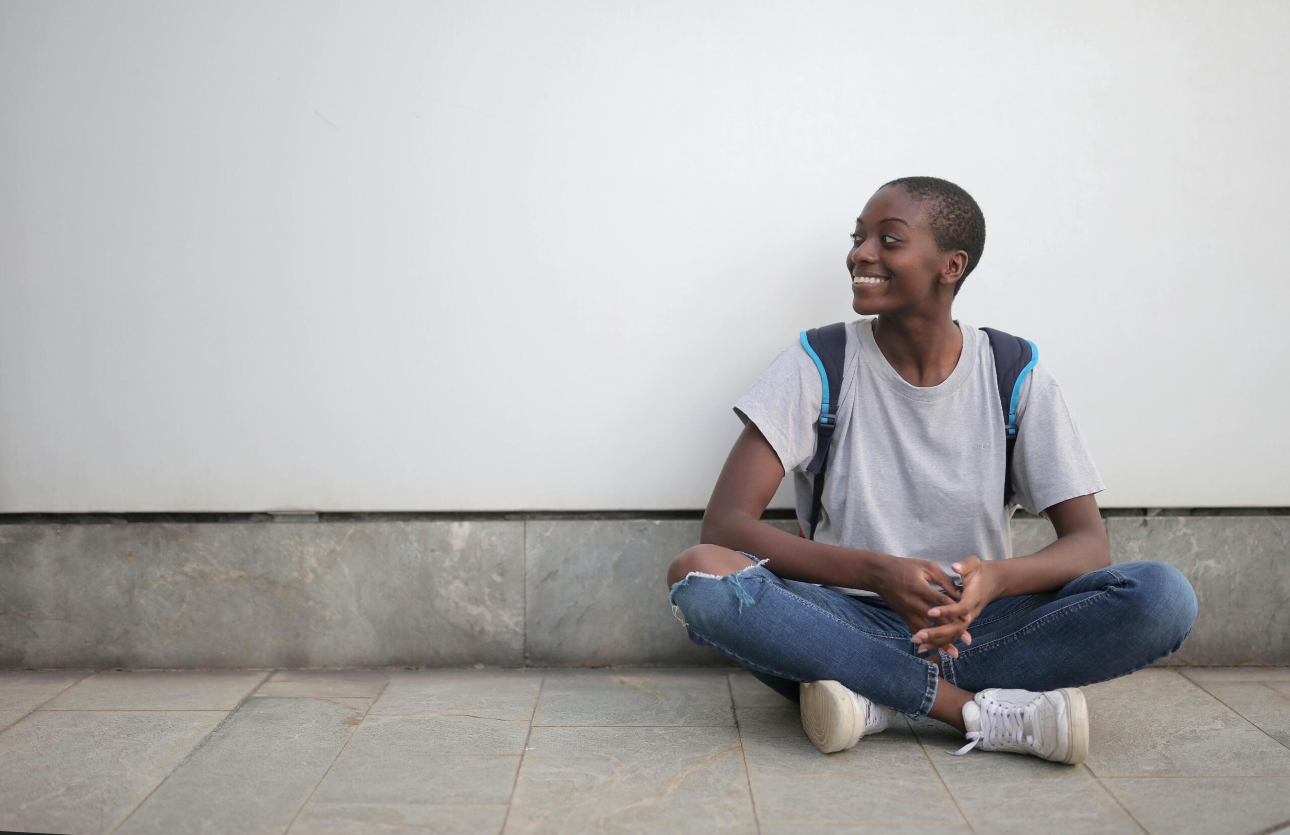 young smiling woman wearing a backpack sitting on the ground in front of a wall