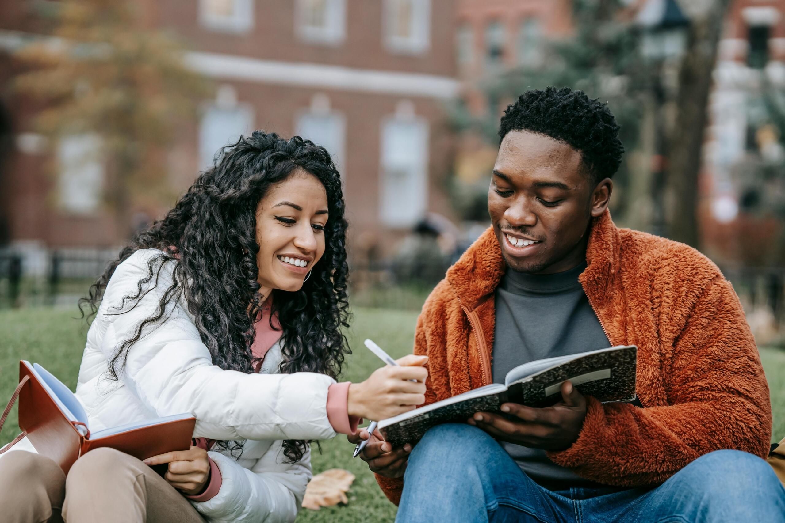 a young woman writing in a young man's notebook as they sit outdoors