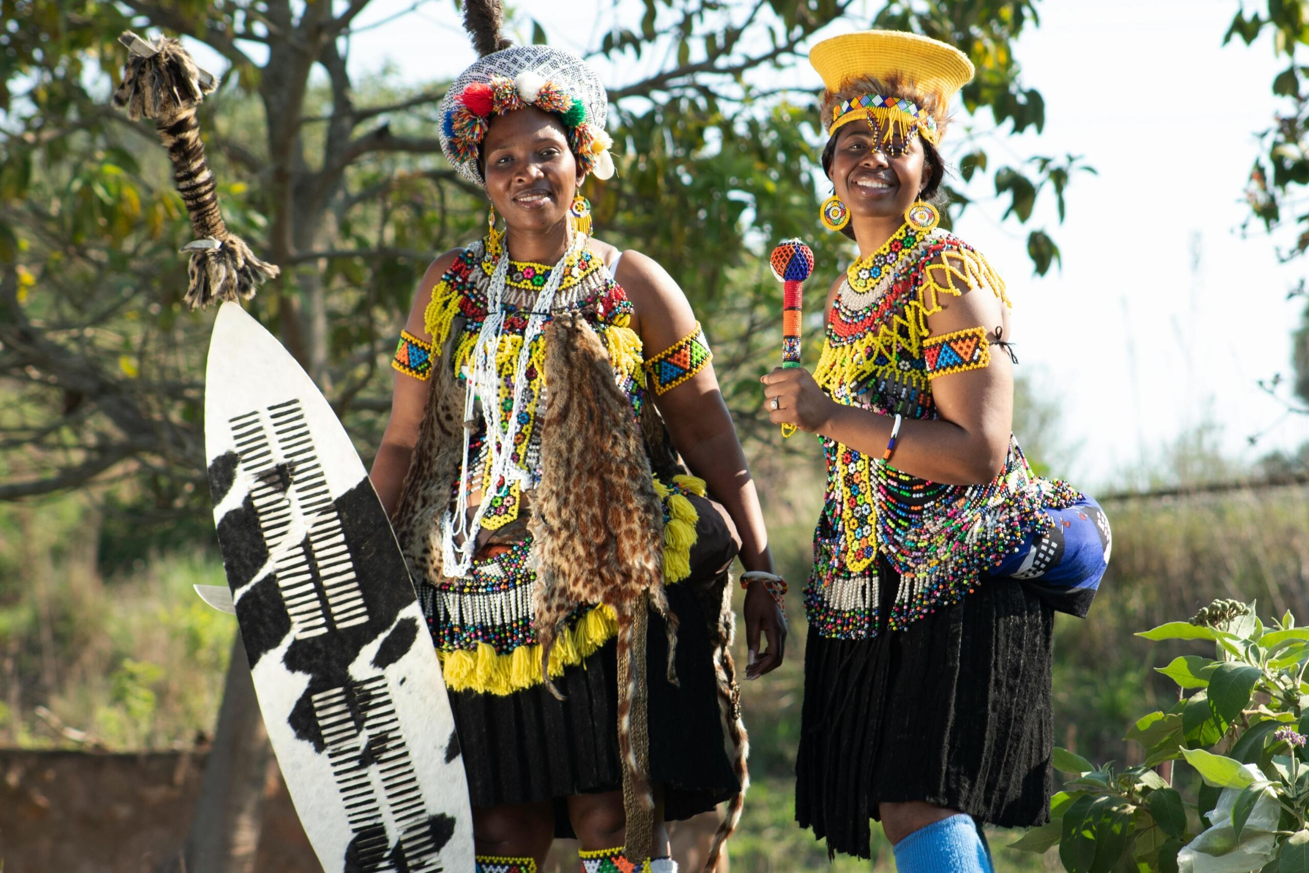two women wearing traditional clothes with beaded belts, necklaces and headgear, holding a shield covered in hide