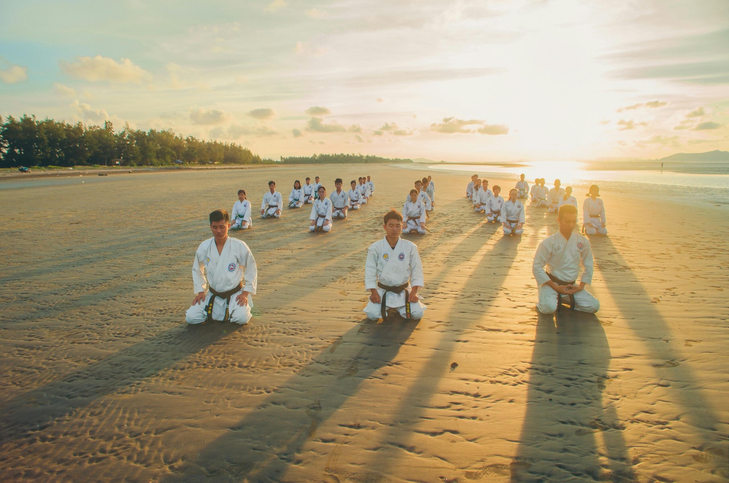 group of martial arts students wearing gis sitting on a beach at sunrise