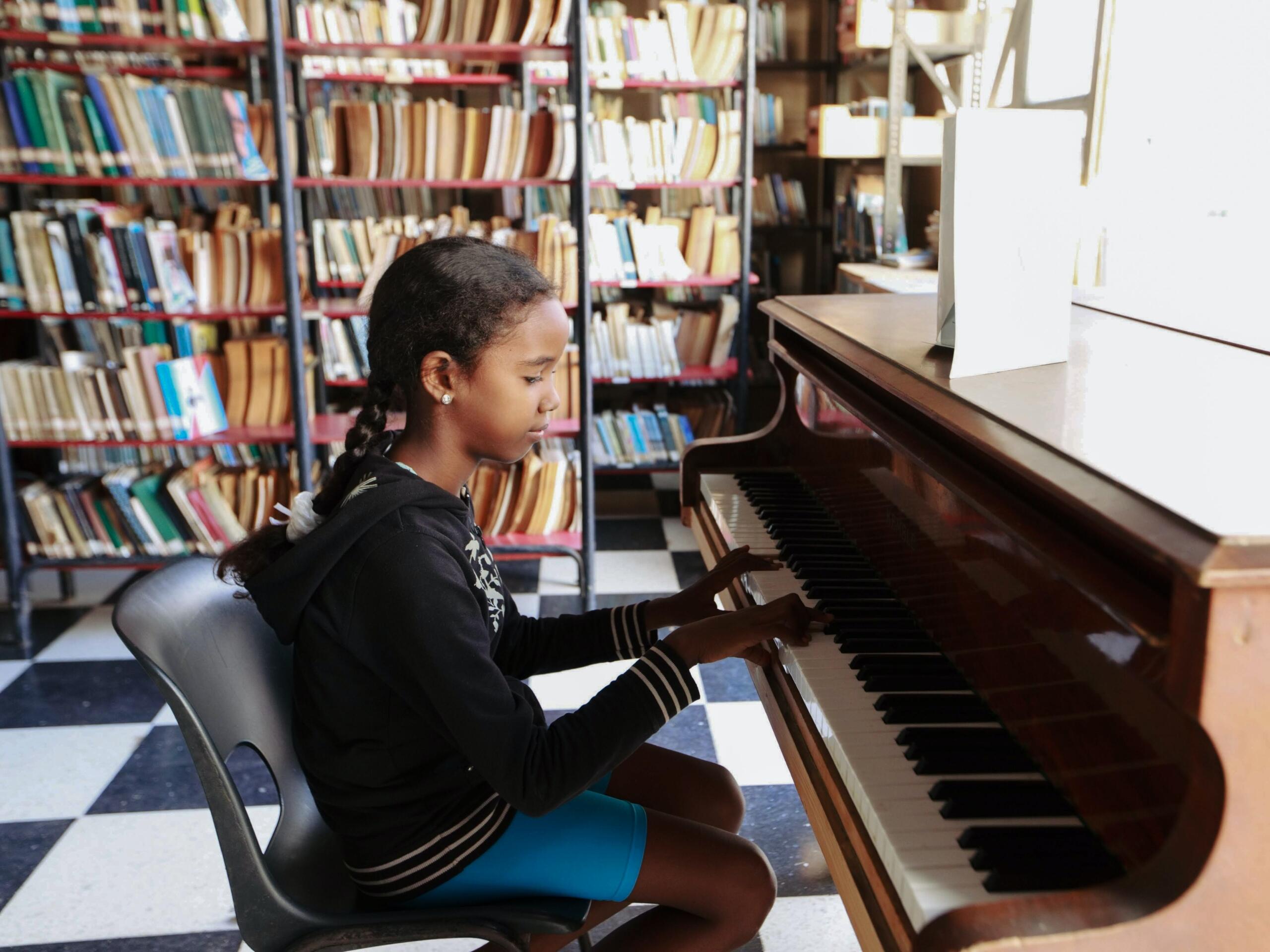 girl reading sheet music and playing brown piano in front of bookshelves