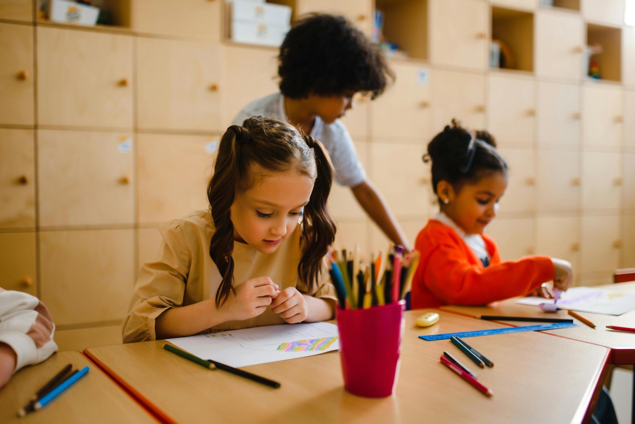 young girls sitting at desks drawing pictures while boy gets up from his seat
