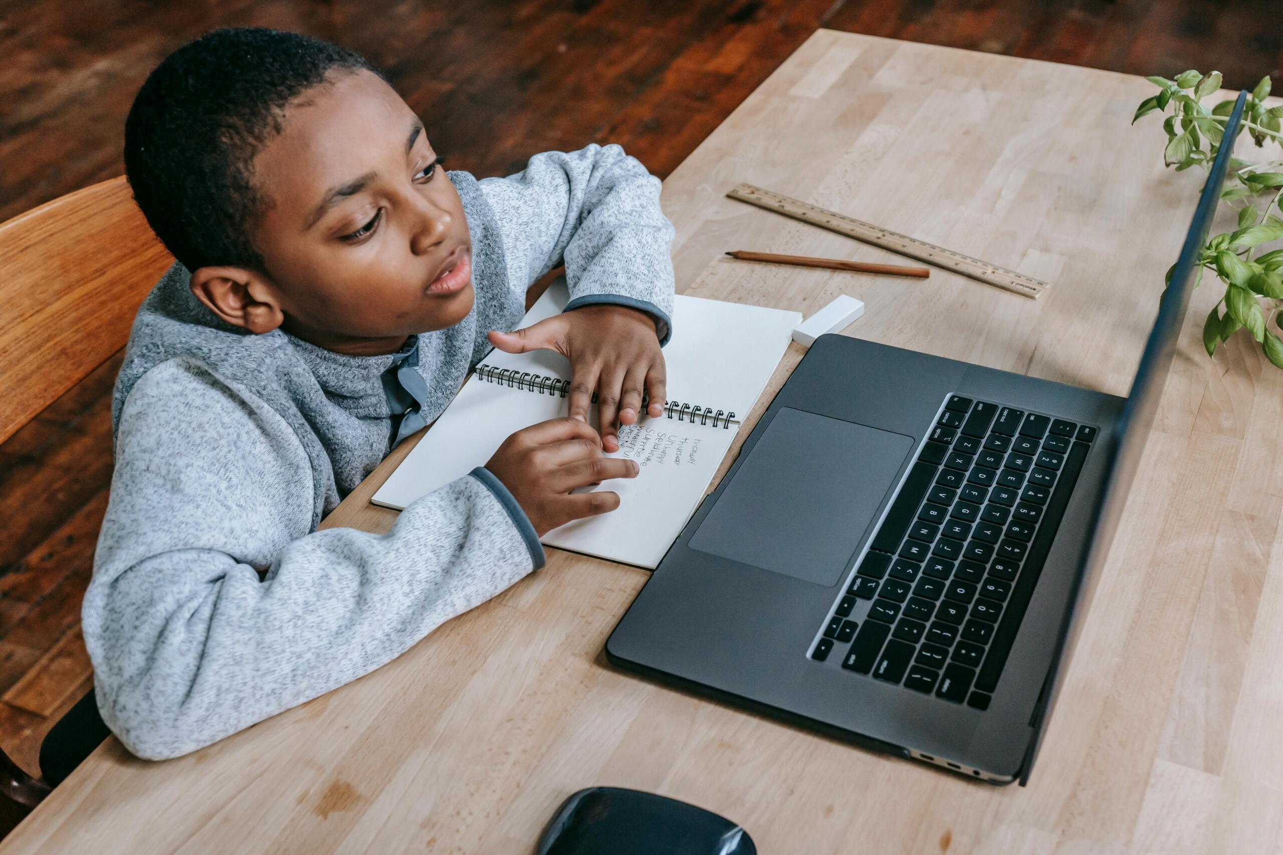 a boy sitting at a desk looking at a laptop screen while working in a notebook
