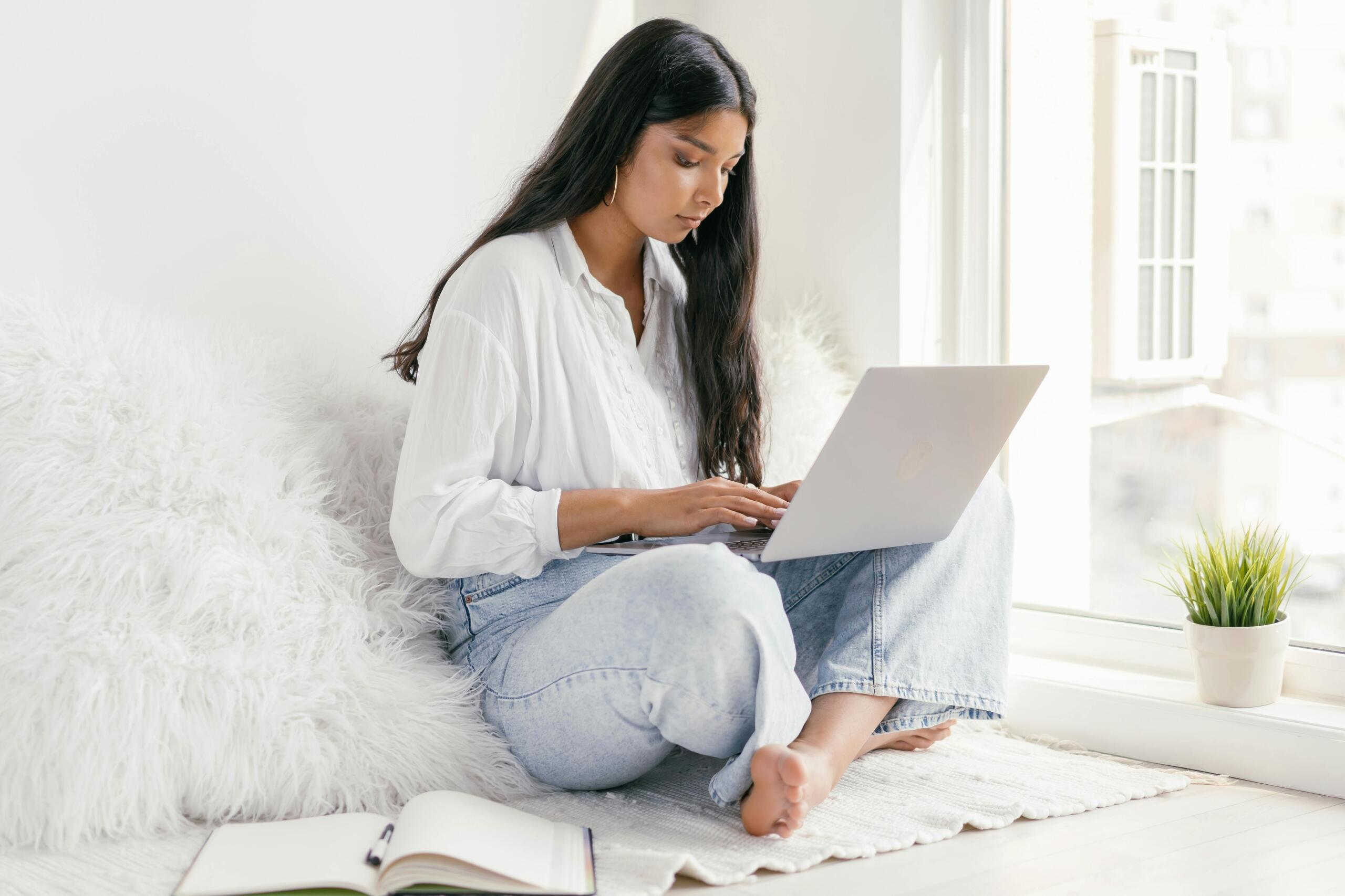 student sitting near window using laptop with an open book nearby