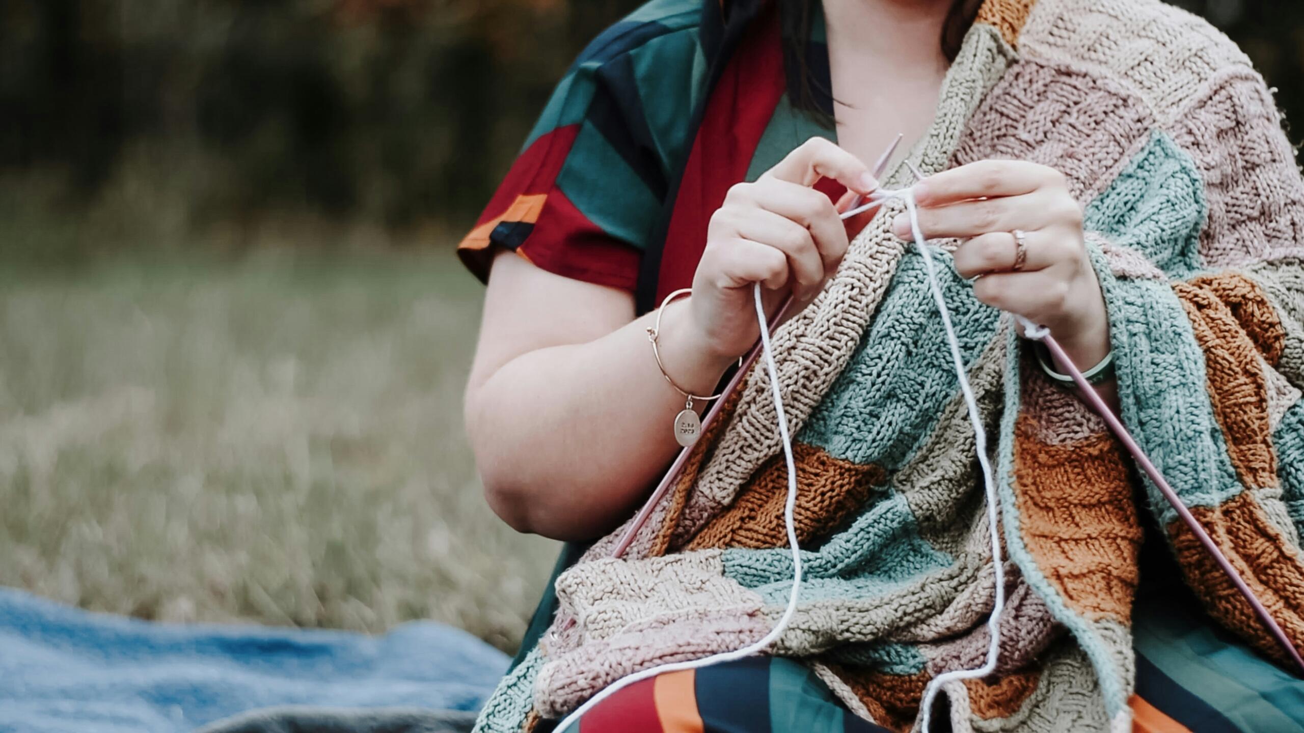 woman sitting outside knitting a blanket that is draped over her shoulders
