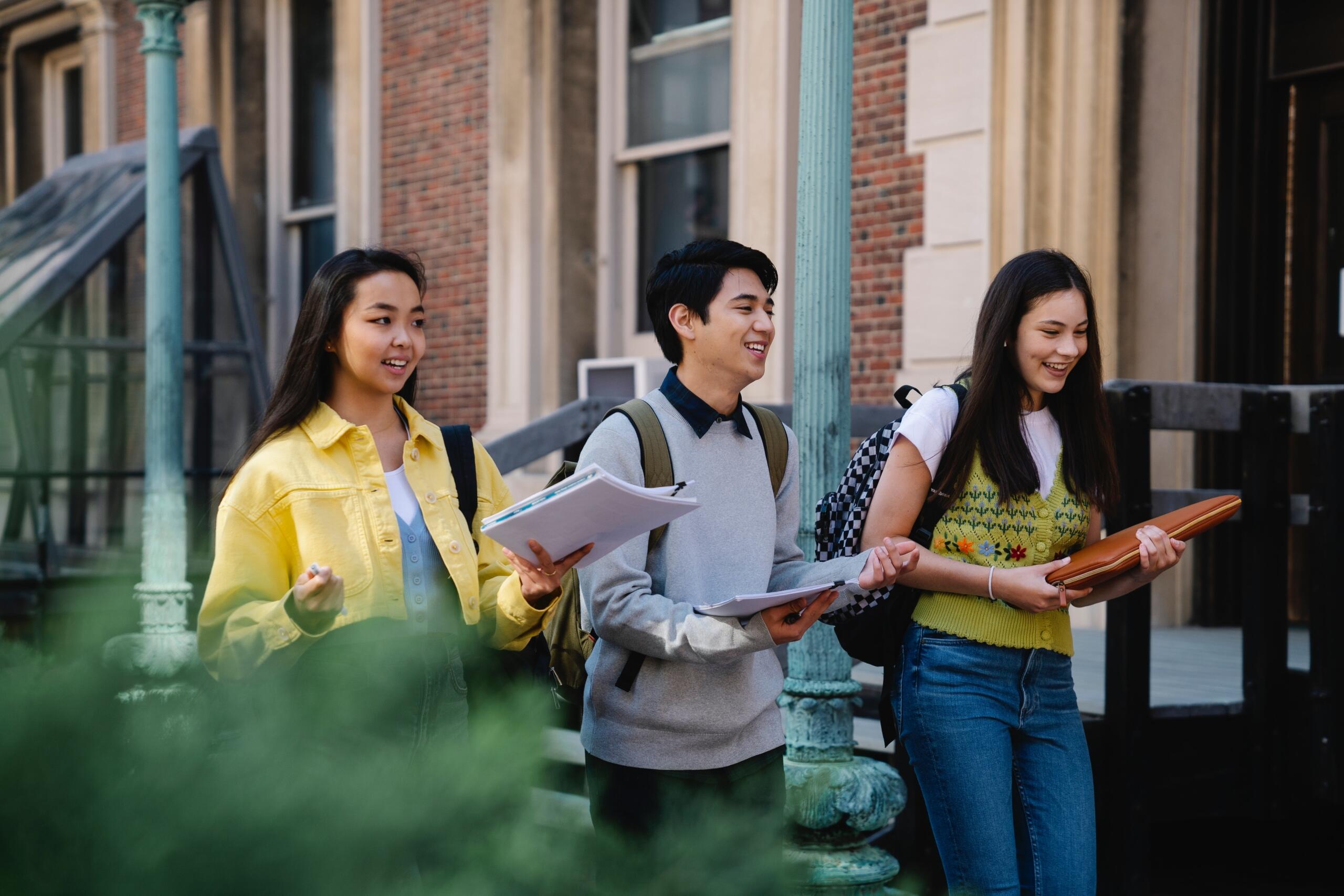 three students holding books and wearing backpacks walking side by side