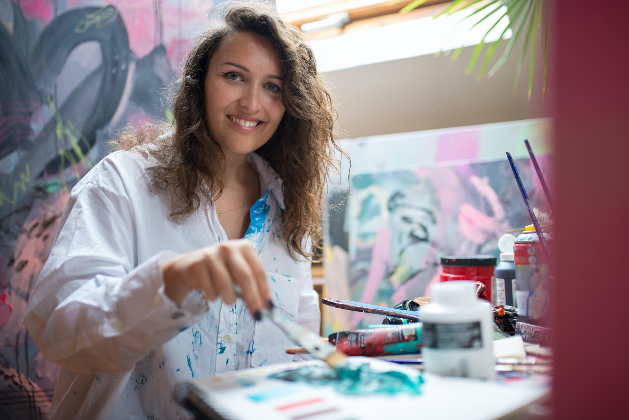 smiling woman holding a paintbrush with multiple painted canvases arranged around her