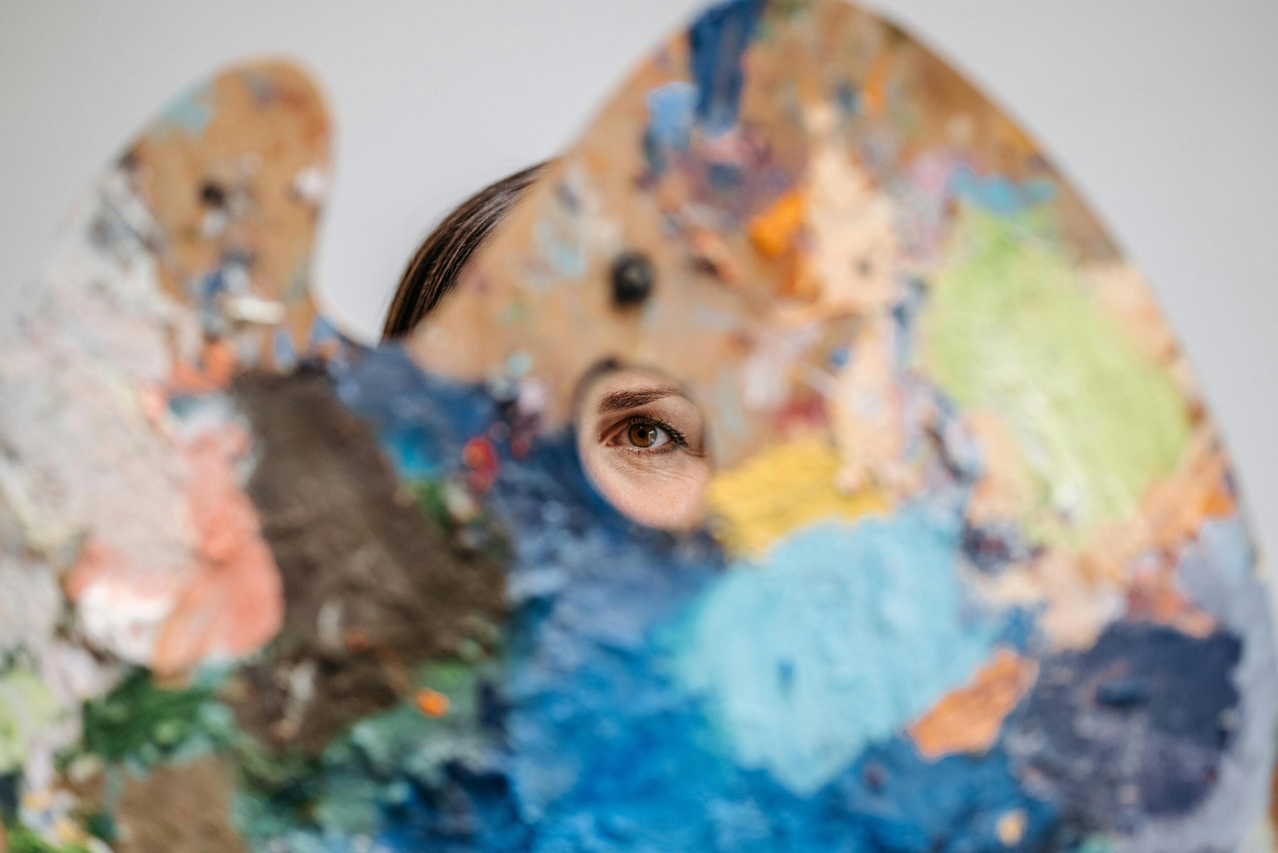 person holding an artist's palette to the camera and peeking through the hold with one eye