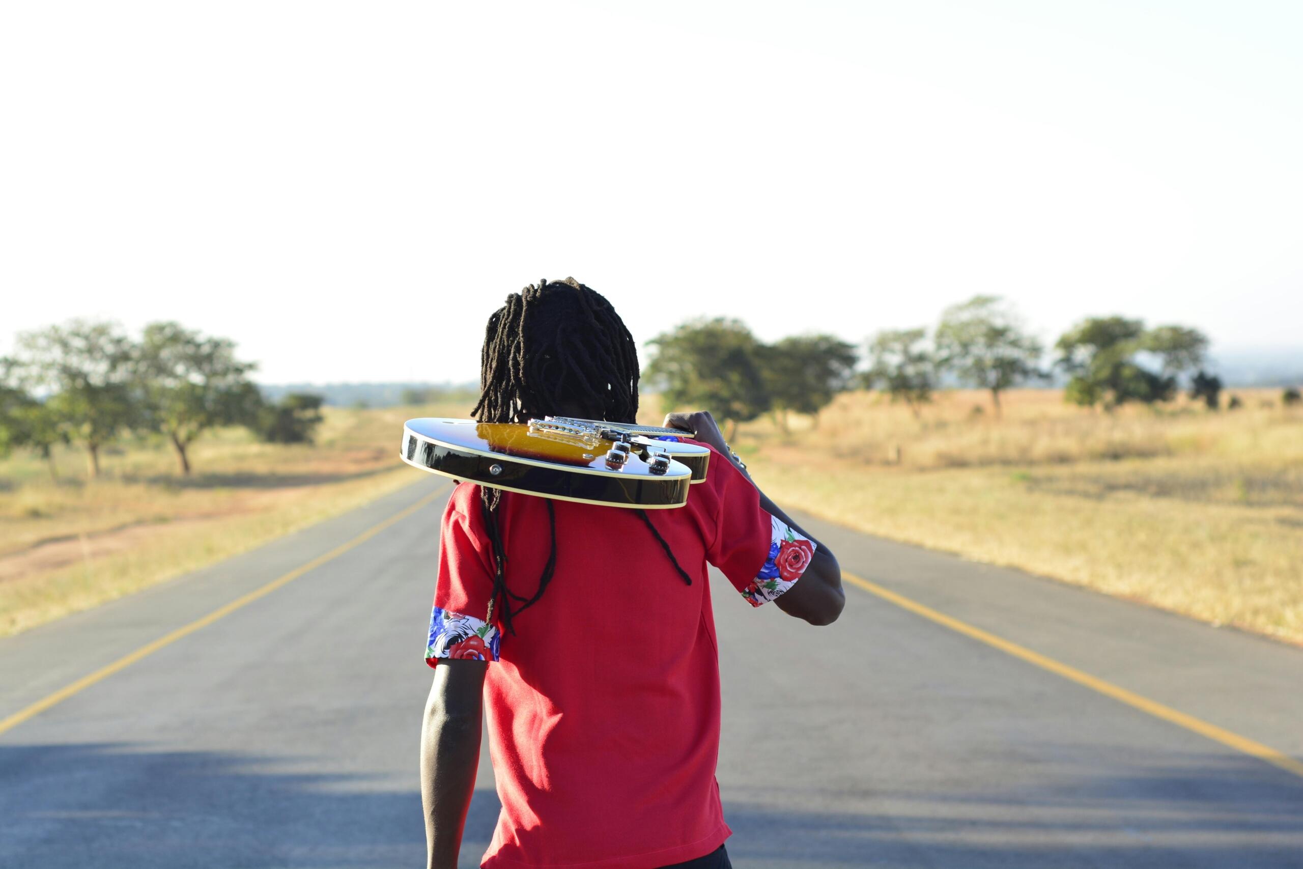 man holding guitar over his shoulder as he walks down a rural raod between grass and trees