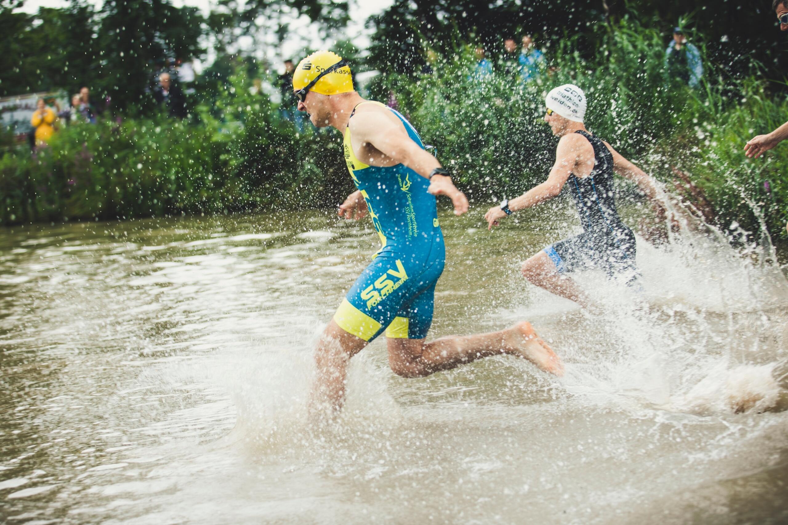 two people in short wetsuits, goggles and swimming caps running into a lake